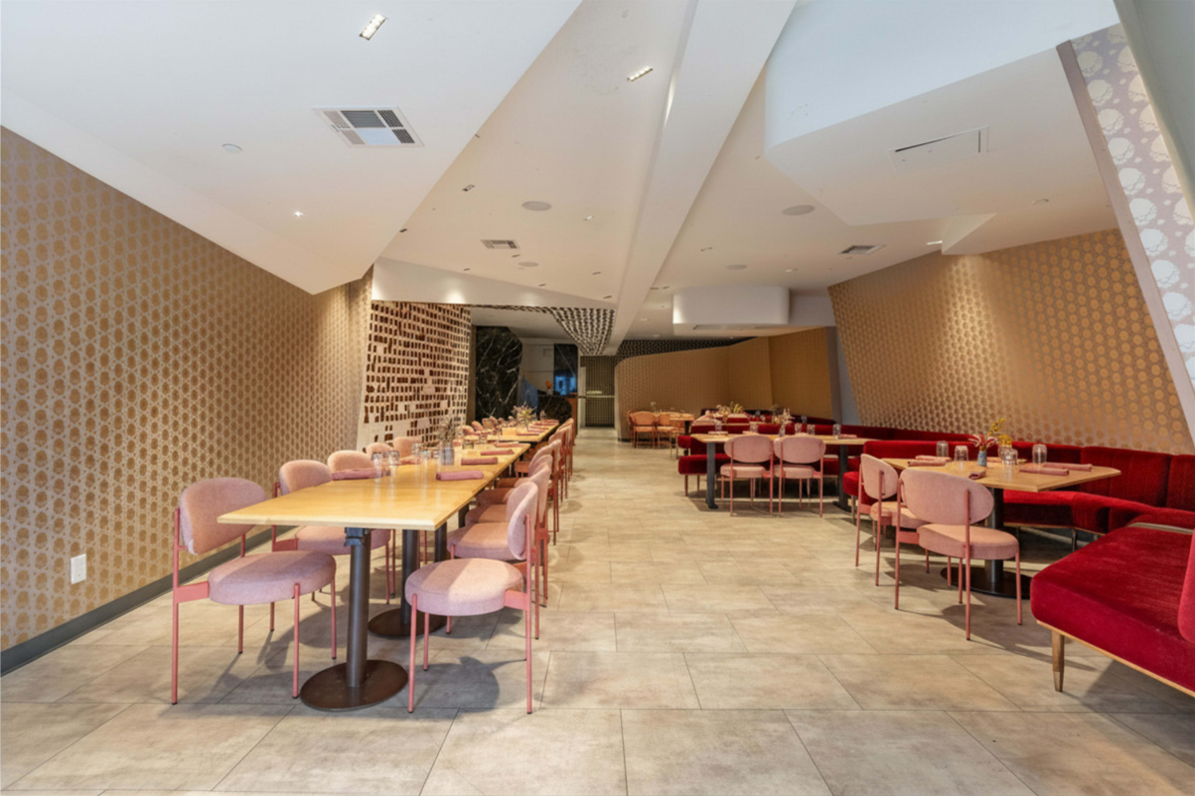 The image shows a modern restaurant interior with rows of tables and chairs arranged on either side, featuring a mix of pink and red upholstery against patterned walls.