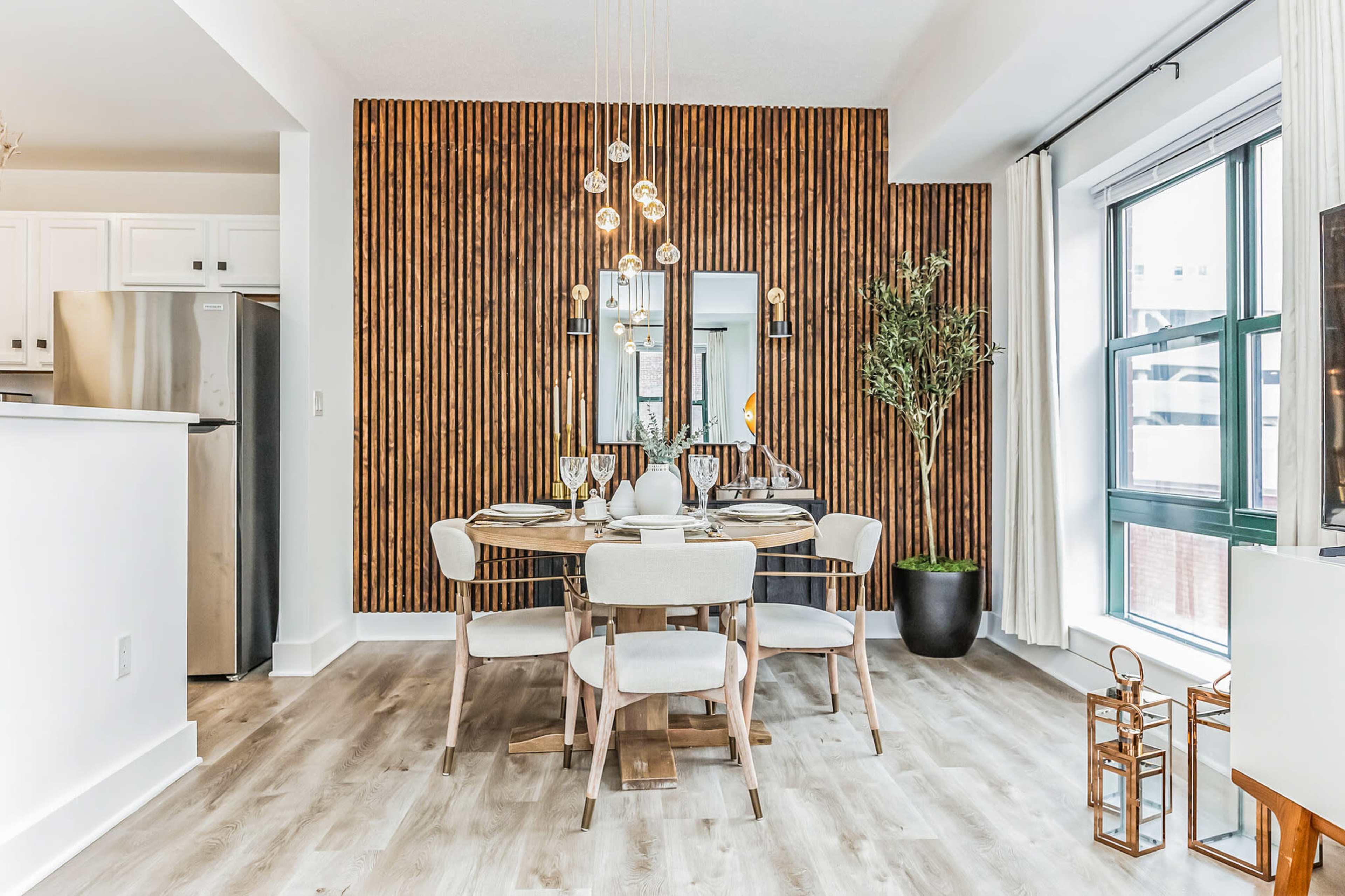 A dining area features a round table set for four, surrounded by chairs, with a wood-paneled accent wall and a modern chandelier above.