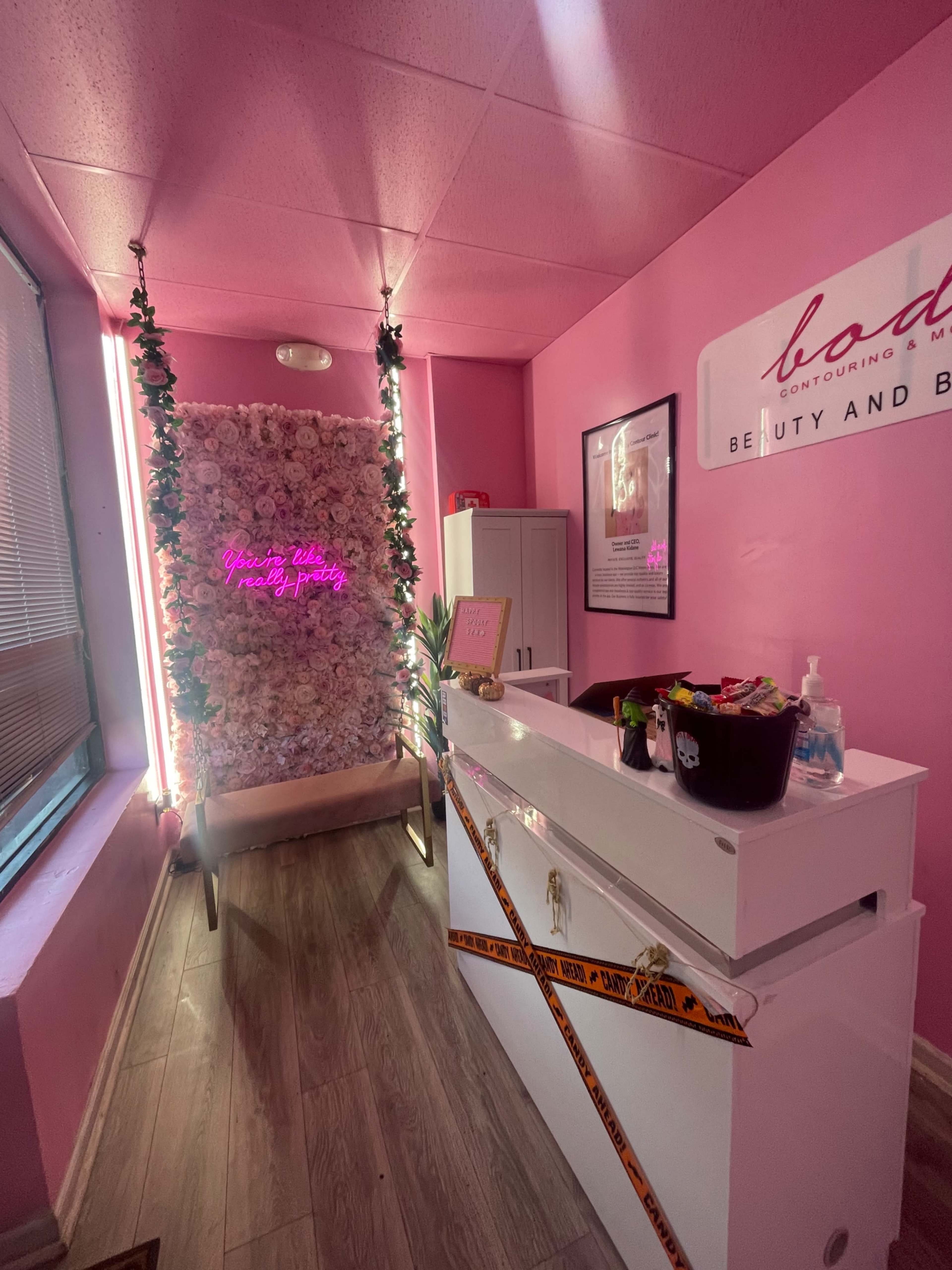 A beauty salon interior featuring a pink-walled reception area with a floral backdrop and a neon sign, along with a white service counter.
