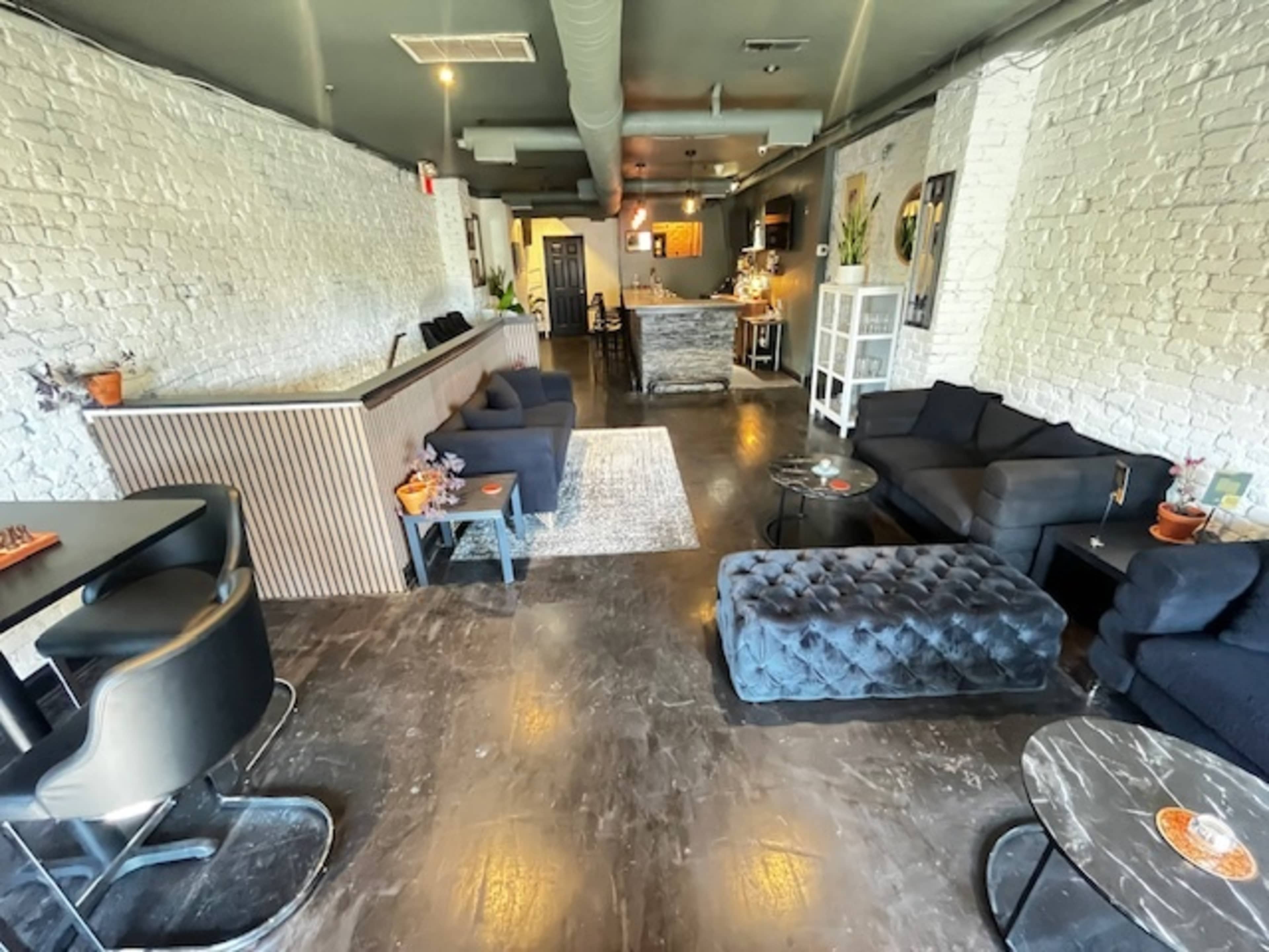 The image shows a stylish interior of a cafe or lounge with brick walls, dark flooring, and various seating arrangements, including sofas and a bar area.