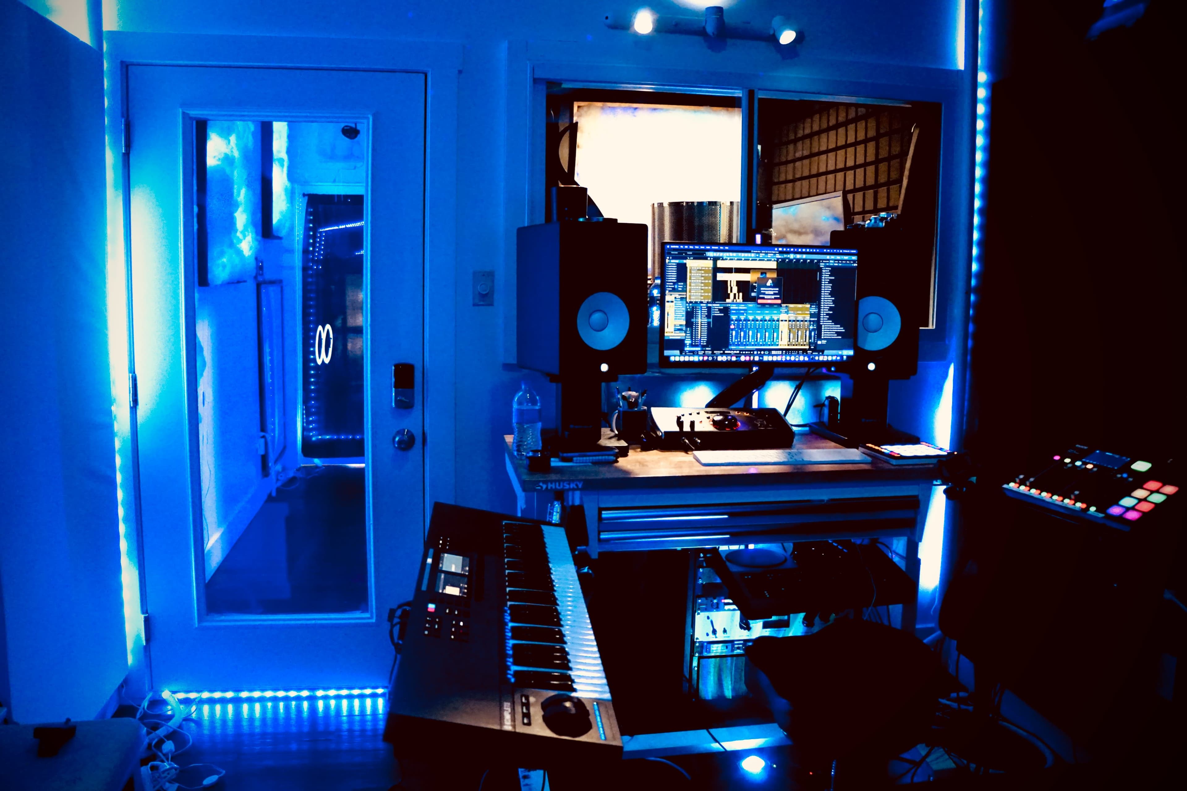 A music production studio featuring a keyboard, computer setup, and LED lighting in blue tones.