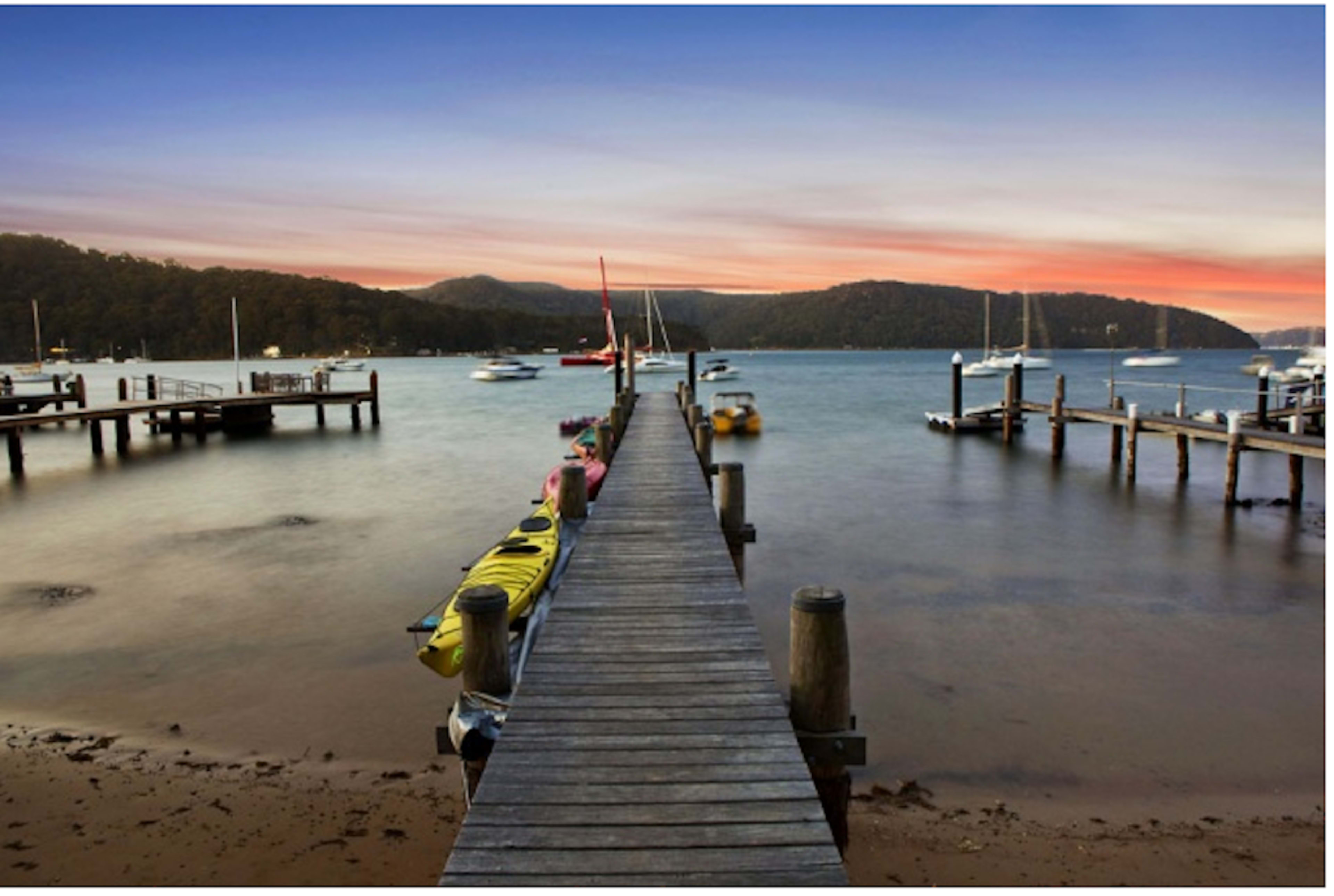 A wooden pier extends into a calm body of water, with anchored boats and hills in the background.