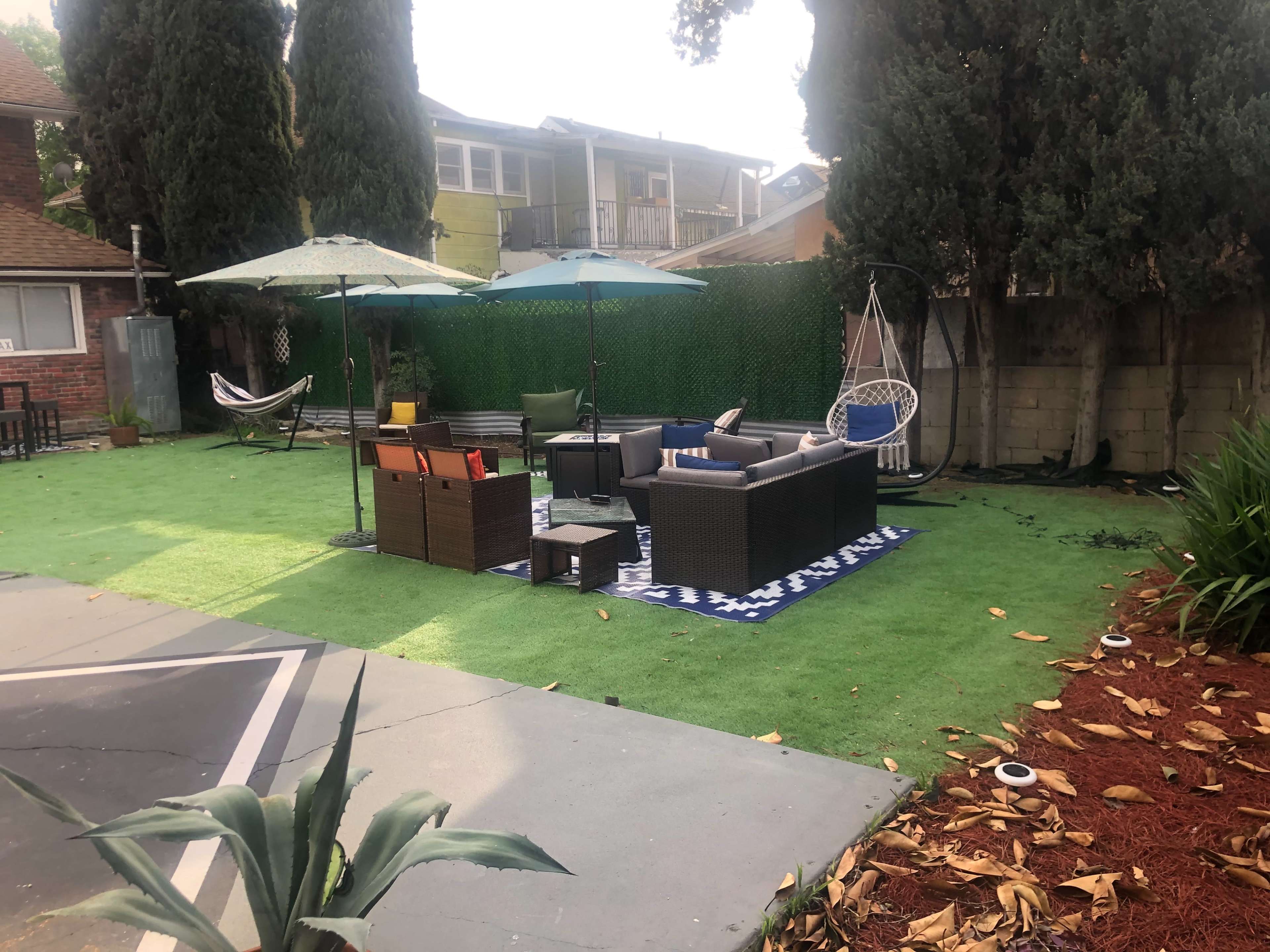 A patio area with green artificial grass, a seating arrangement of dark wicker furniture, two umbrellas, and a hammock in a residential backyard.