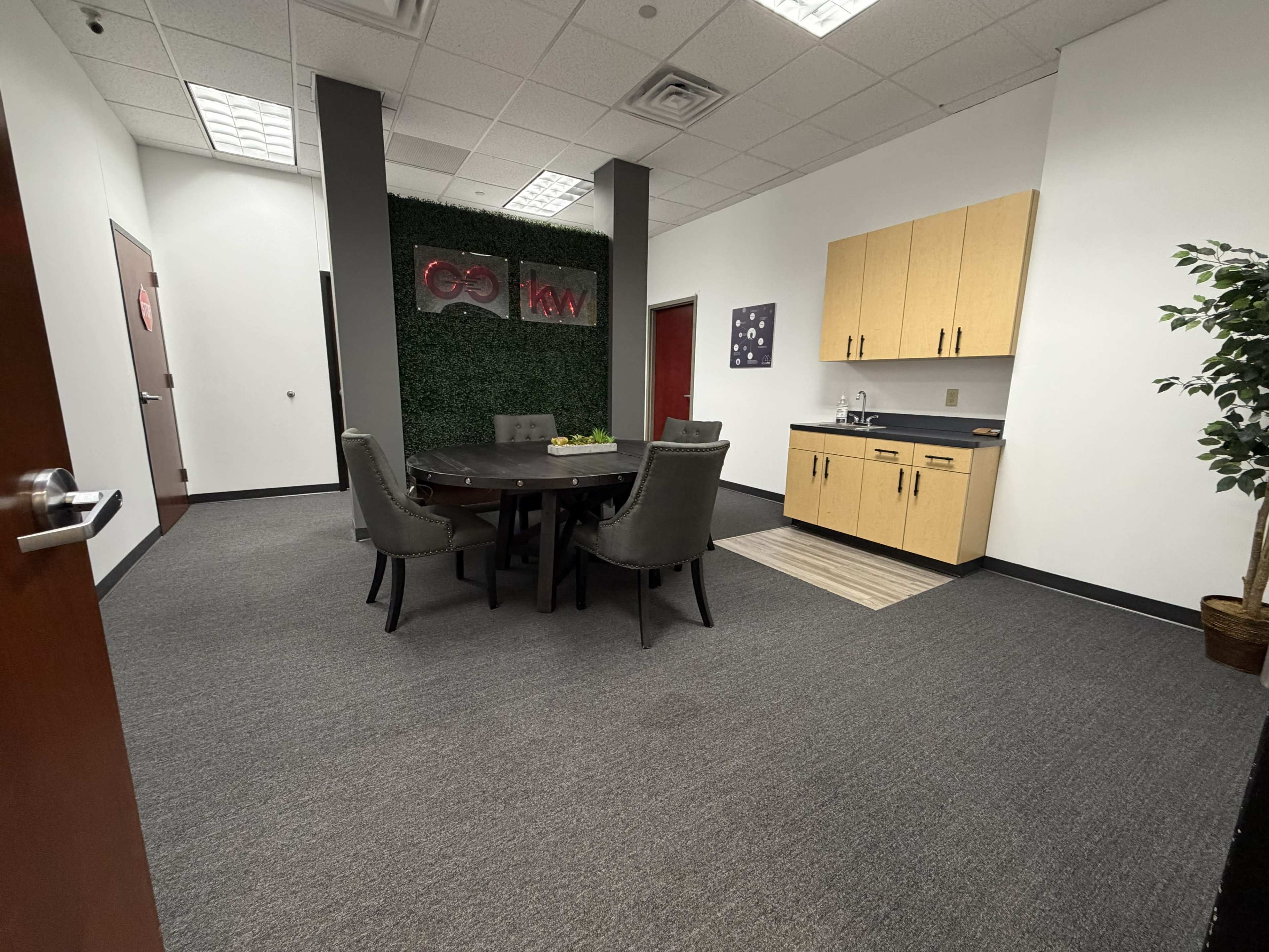 The image shows a meeting room with a round table surrounded by four chairs, a kitchenette along one wall, and a decorative plant in the corner.