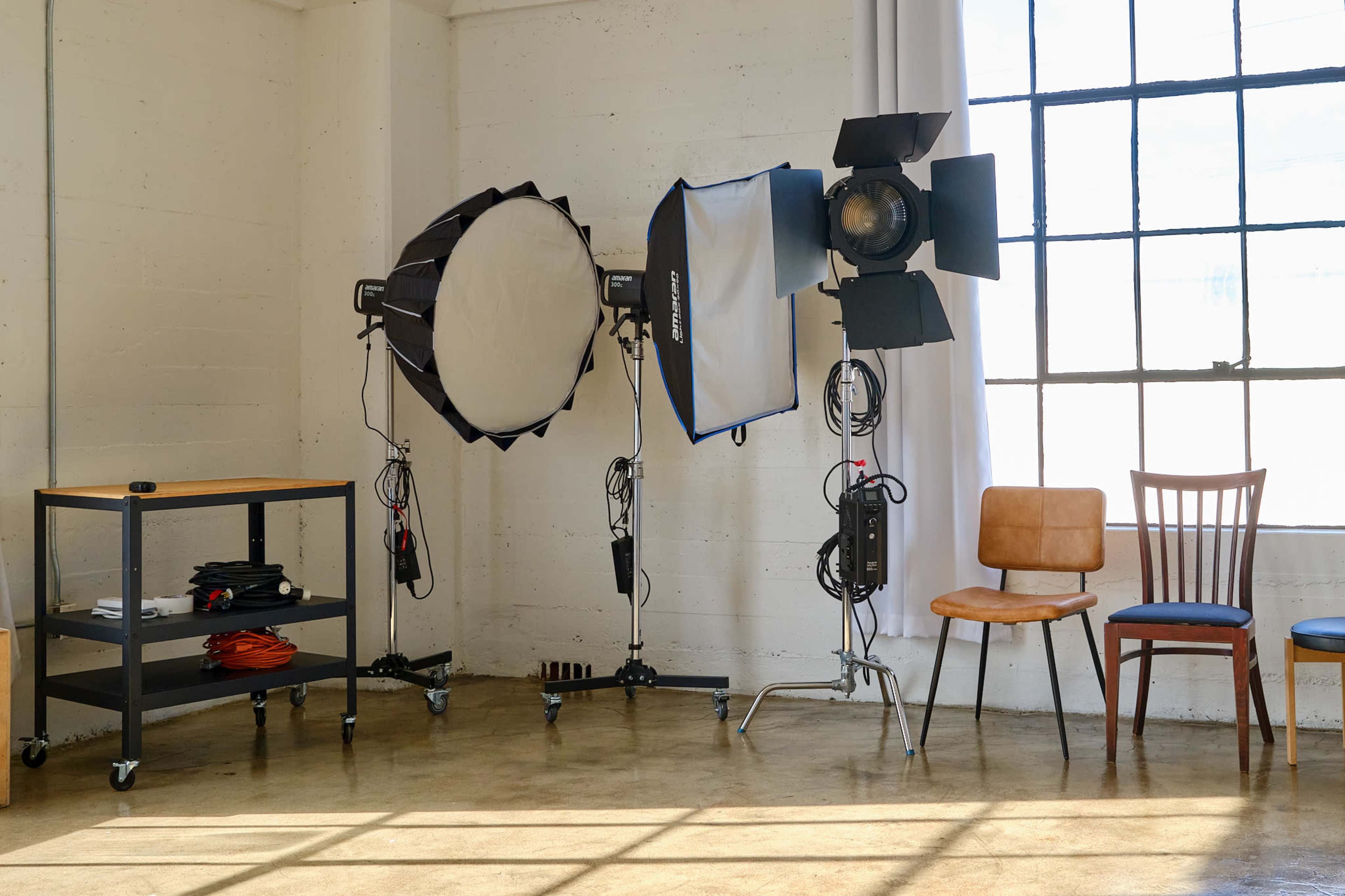 The image shows a well-lit studio space featuring two professional lighting setups and a wooden cart with equipment, alongside a brown chair and a wooden chair near a large window.