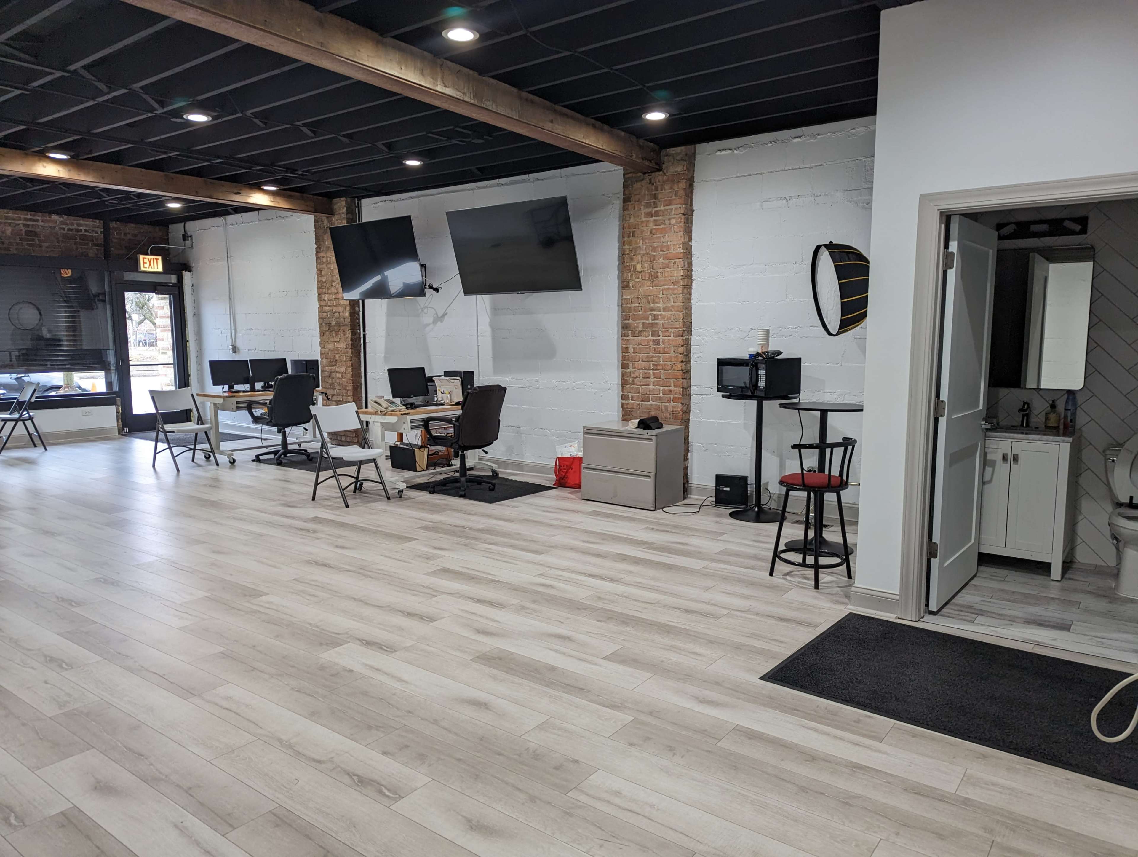 A spacious office area with several workstations, two large screens on the wall, and a small kitchenette near a bathroom door.