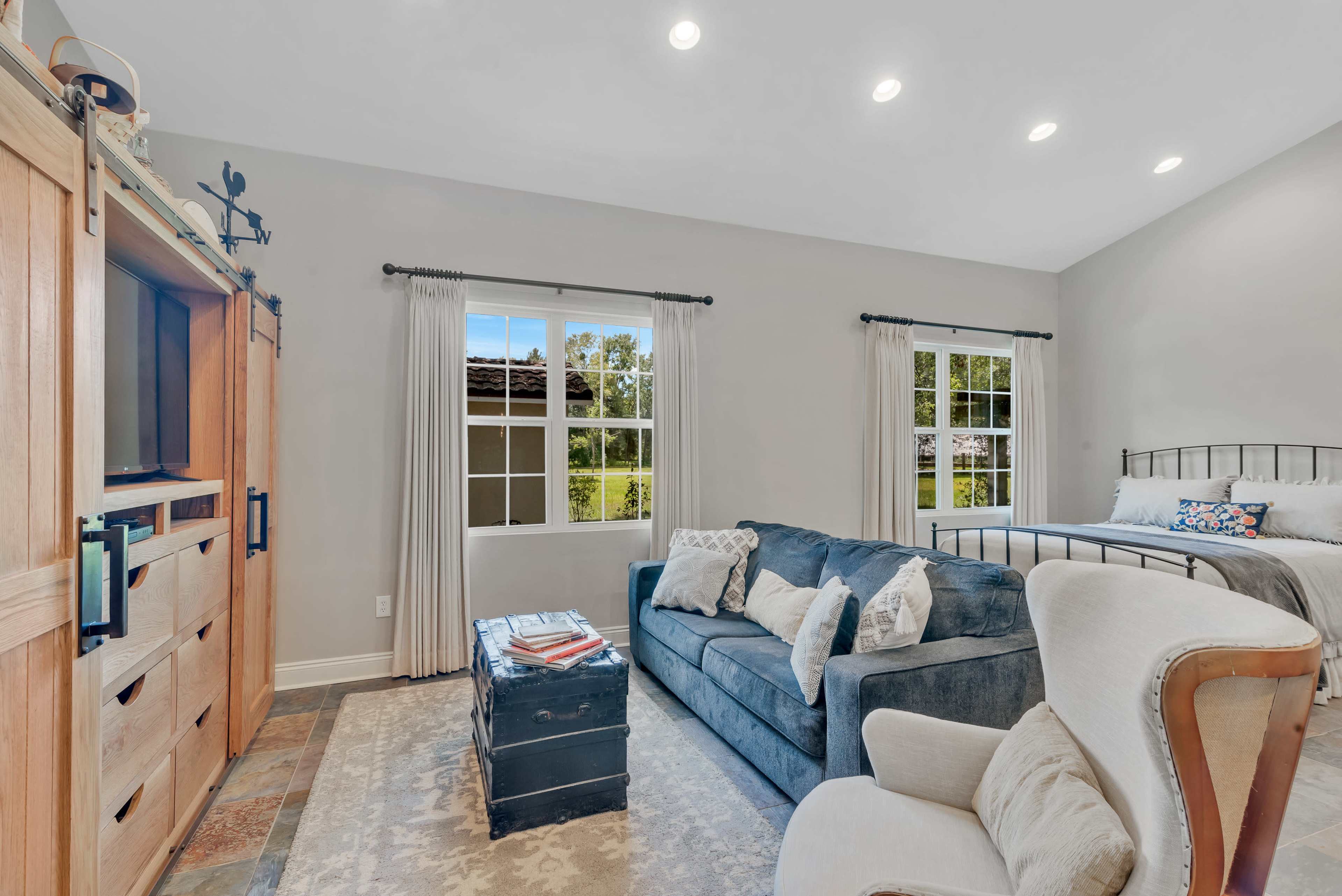The room features a sofa and armchair, a wooden entertainment unit, and large windows overlooking greenery.