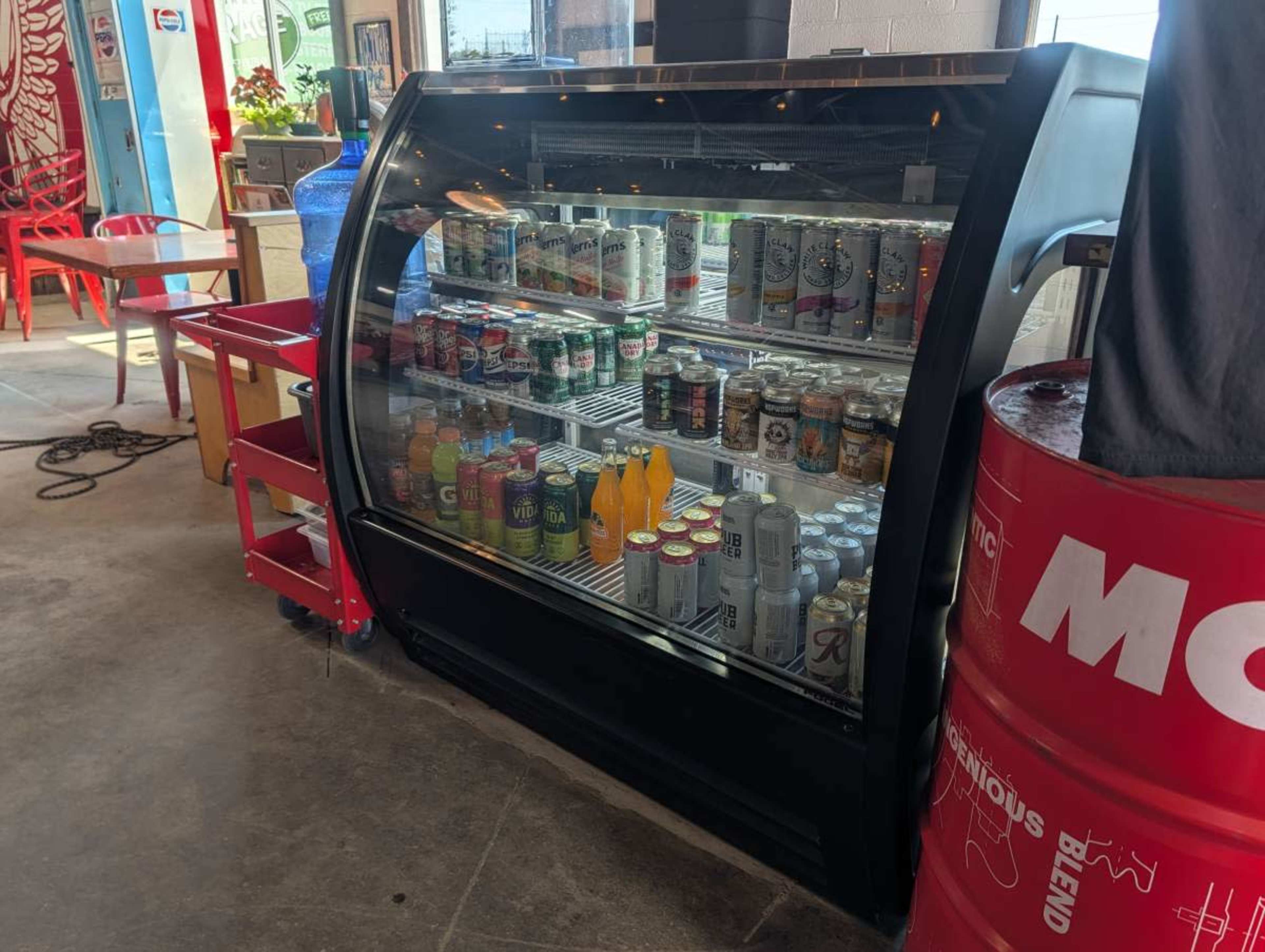 The image shows a glass front beverage cooler filled with various cans and bottles in a casual dining space.