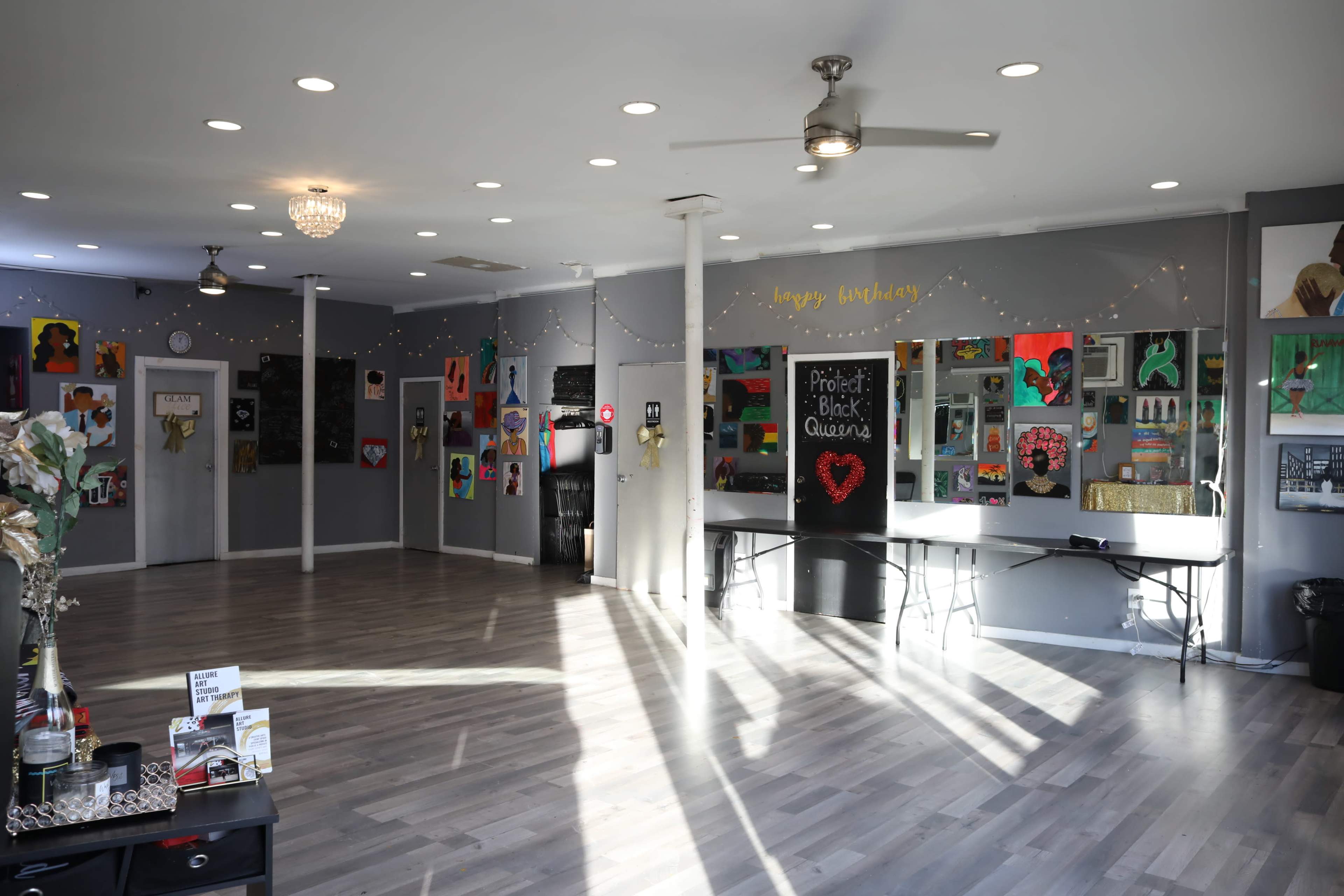 The interior of a spacious art studio featuring various colorful paintings on the walls, tables set up for display, and bright overhead lighting.