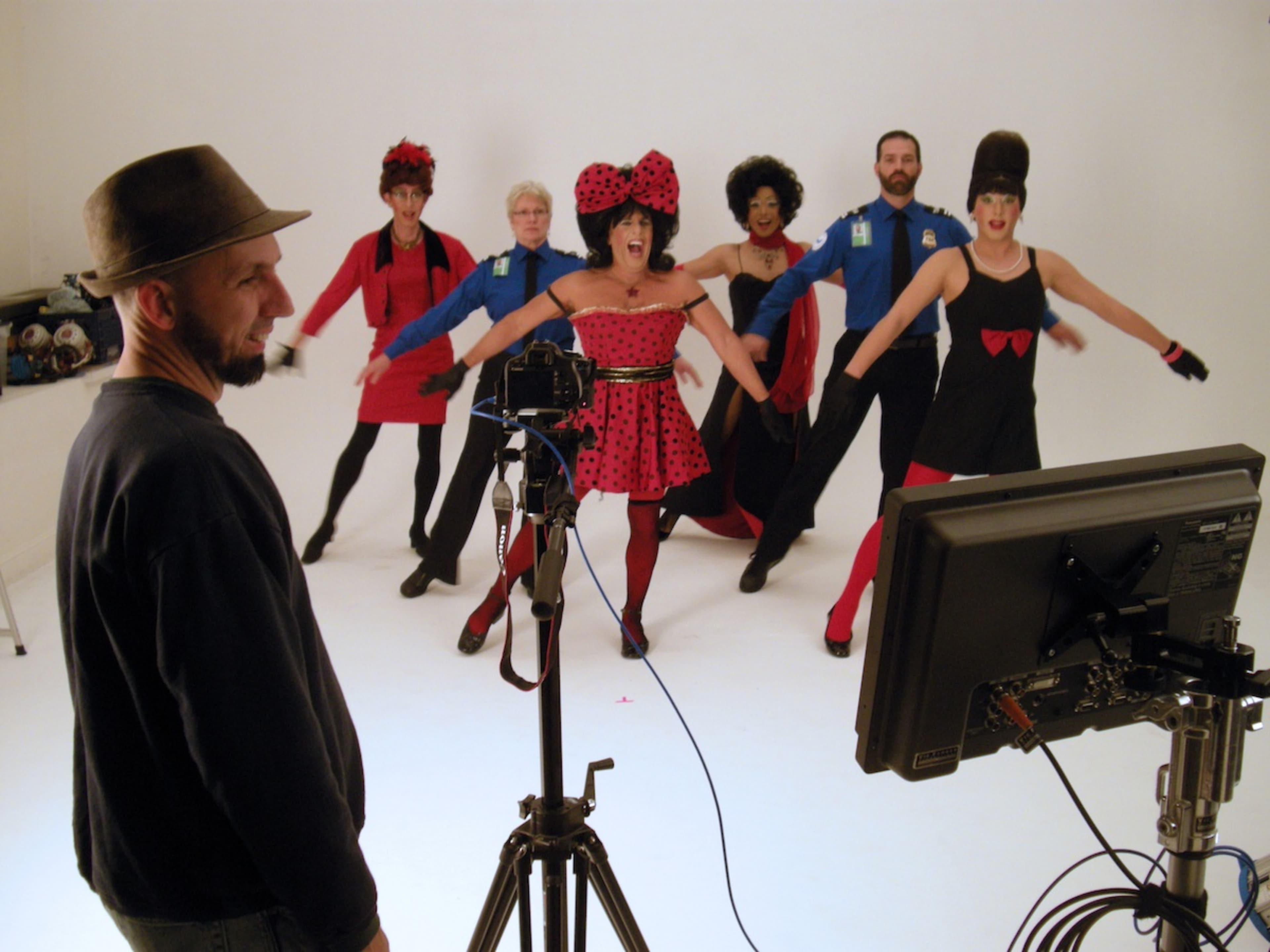 A group of performers in colorful costumes and wigs poses energetically in front of a camera while a man in a hat observes from behind.