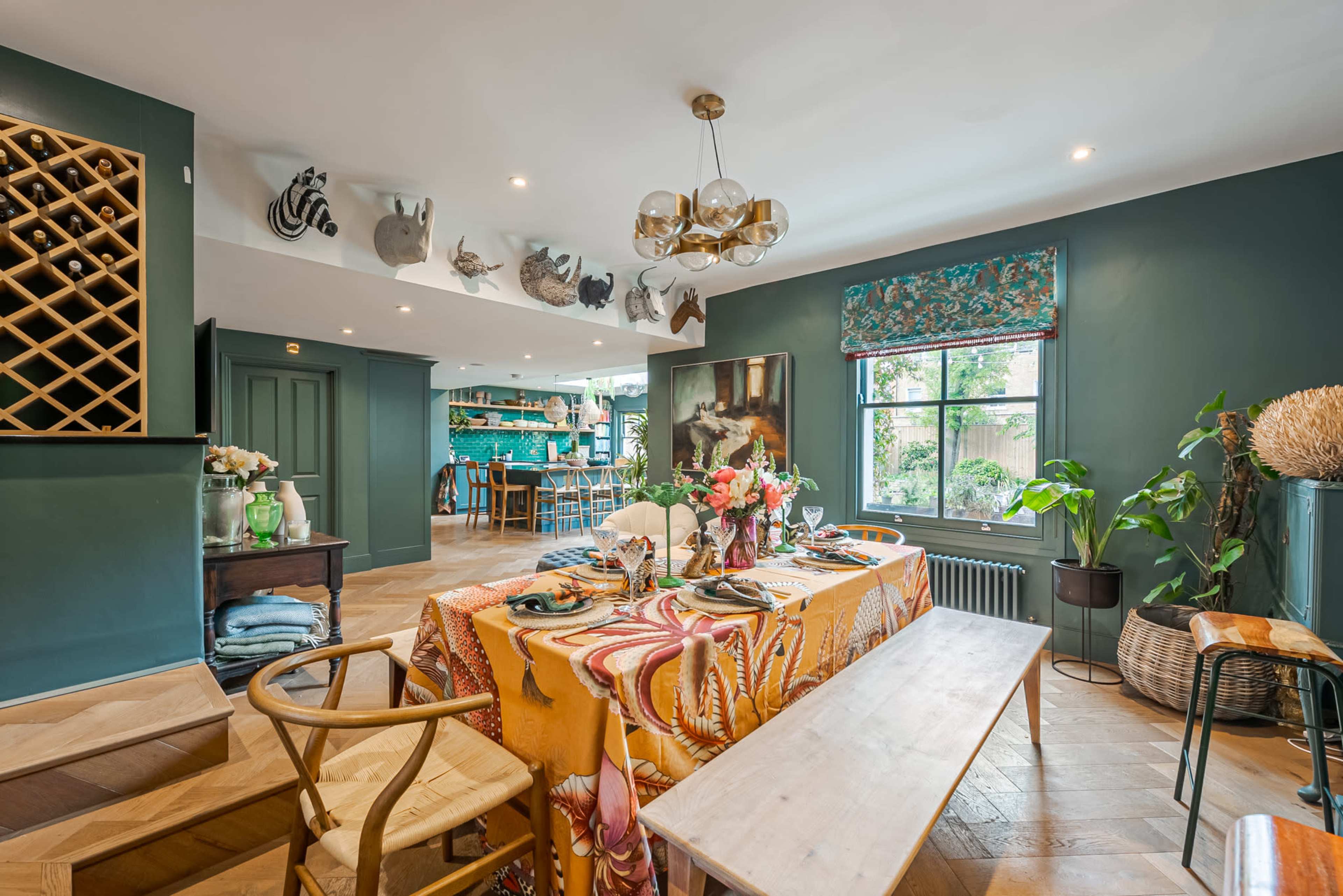 The image shows a dining area featuring a colorful table setting with a patterned tablecloth, surrounded by chairs and leading to a kitchen area in the background.