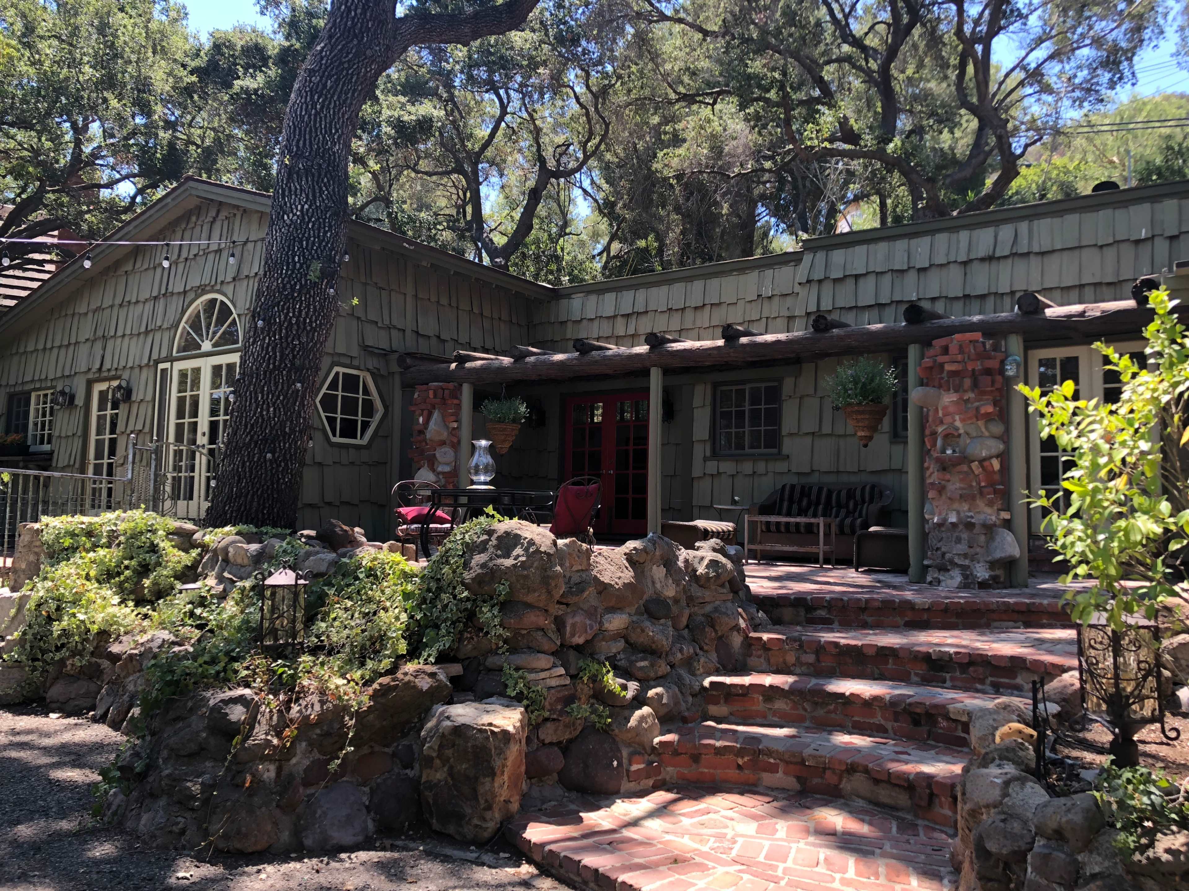 A rustic house with a stone path and a porch, surrounded by greenery and trees.