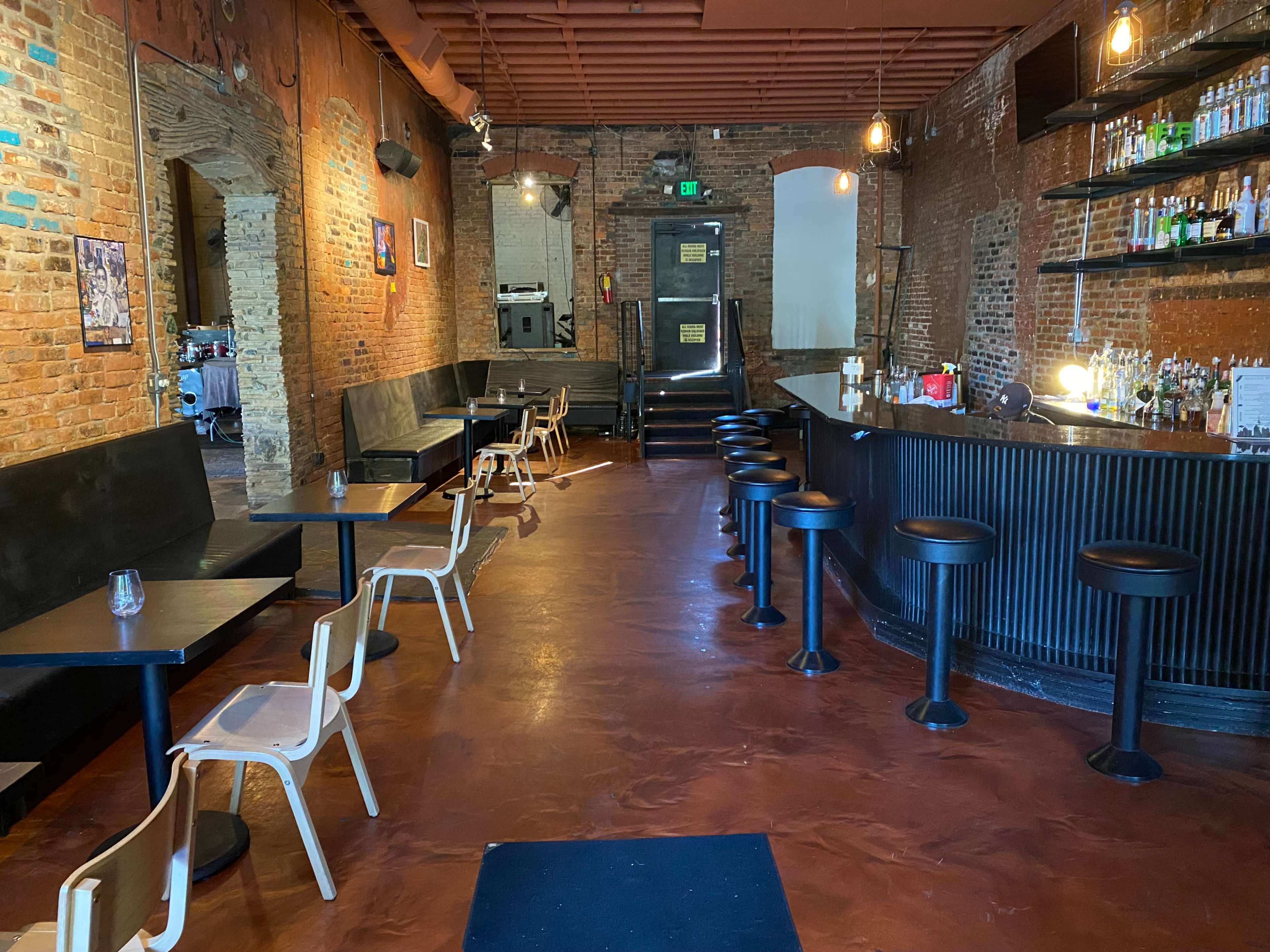 A dimly lit restaurant interior with brick walls, a bar on the right, and several tables and chairs lining the left side.