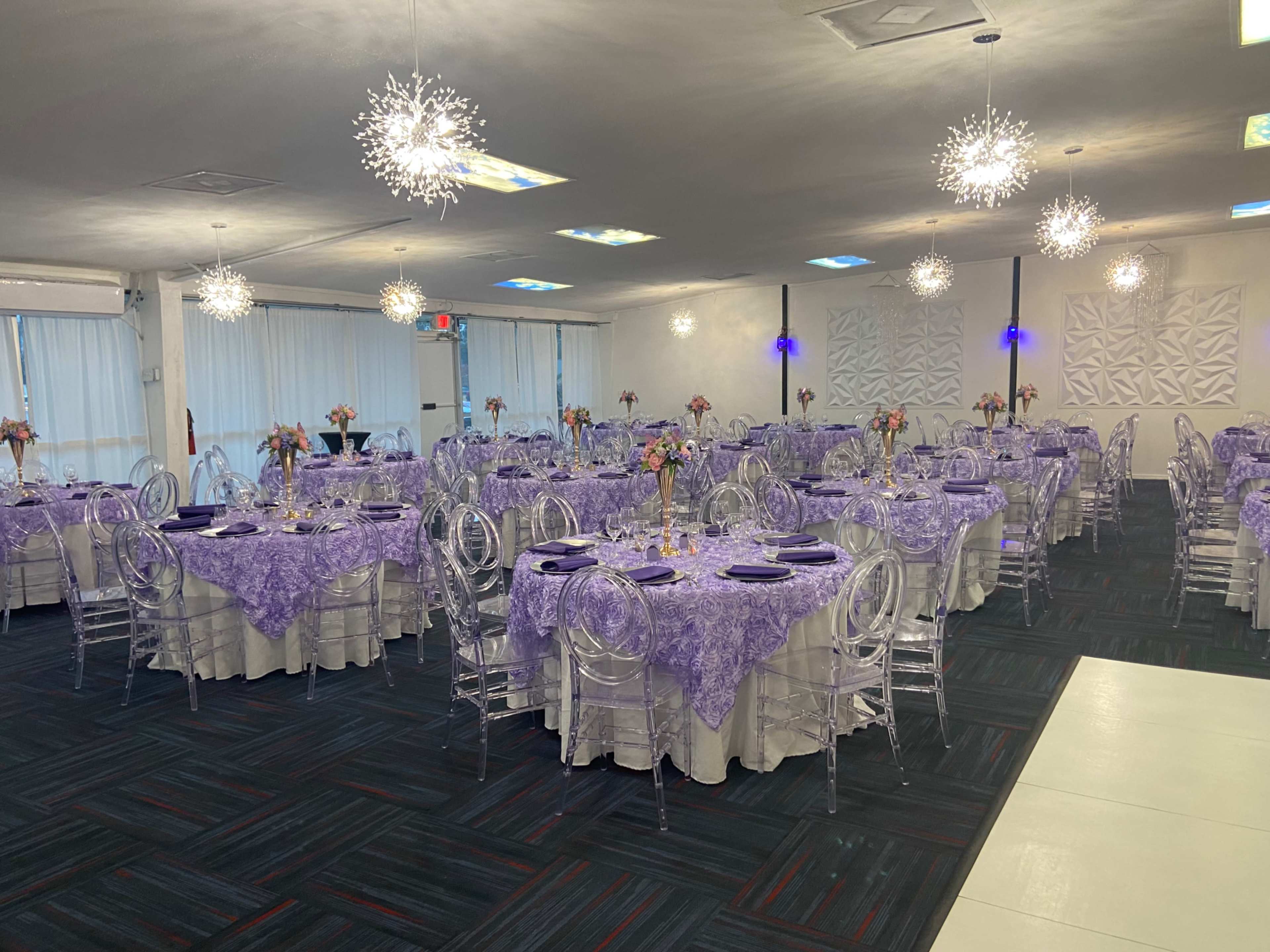 The image shows a banquet hall set up with round tables covered in lavender tablecloths, each adorned with floral centerpieces and surrounded by clear acrylic chairs.