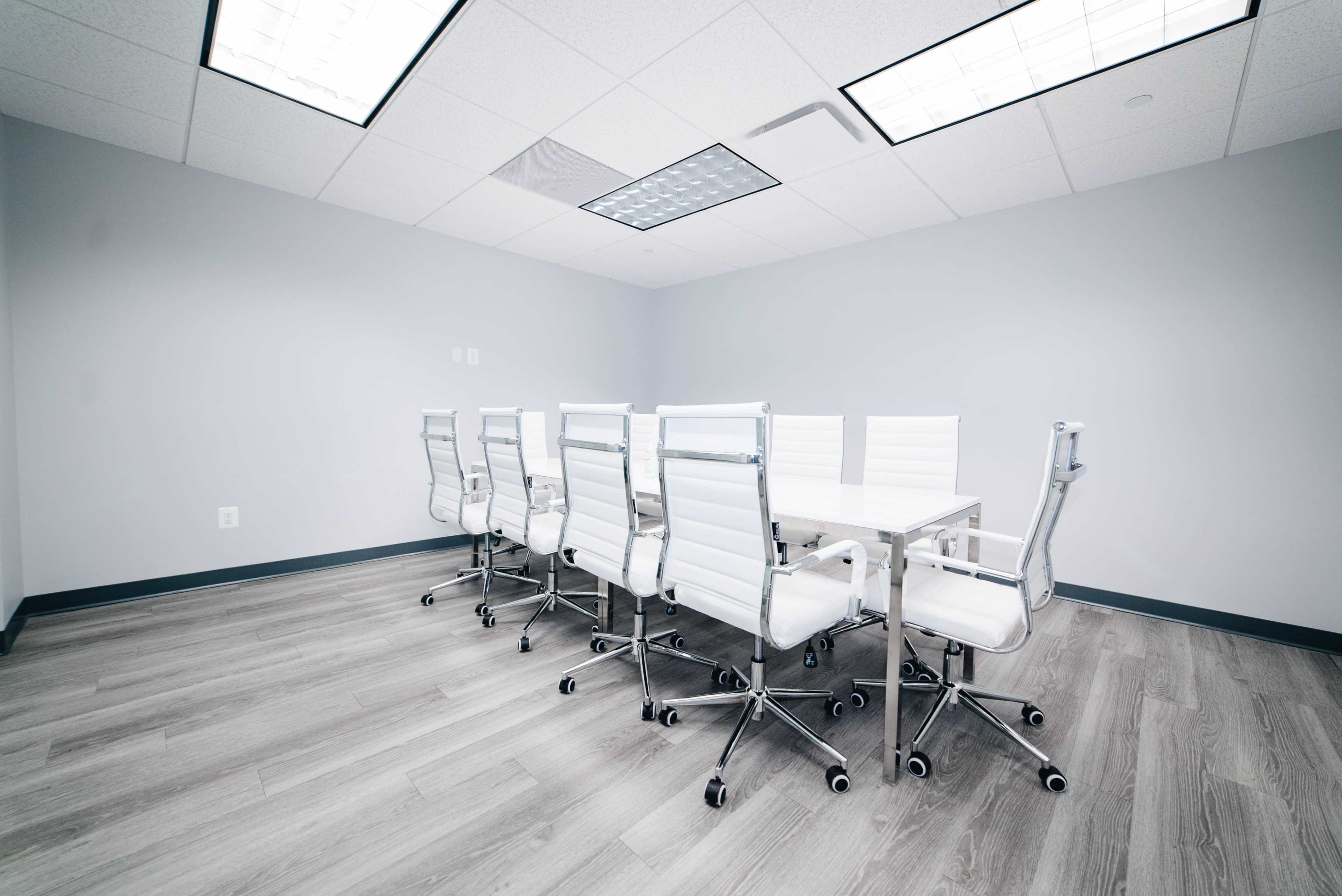 A modern conference room features a white table surrounded by seven sleek, white office chairs on a hardwood floor.