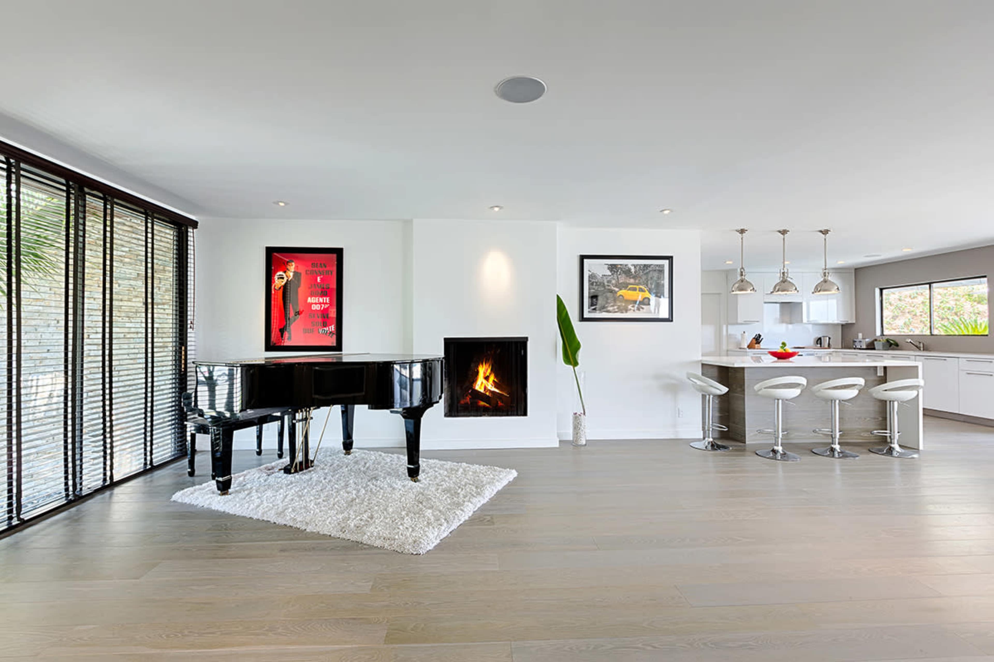 A modern living space features a black grand piano on a white rug, a fireplace, and a kitchen bar with stools.