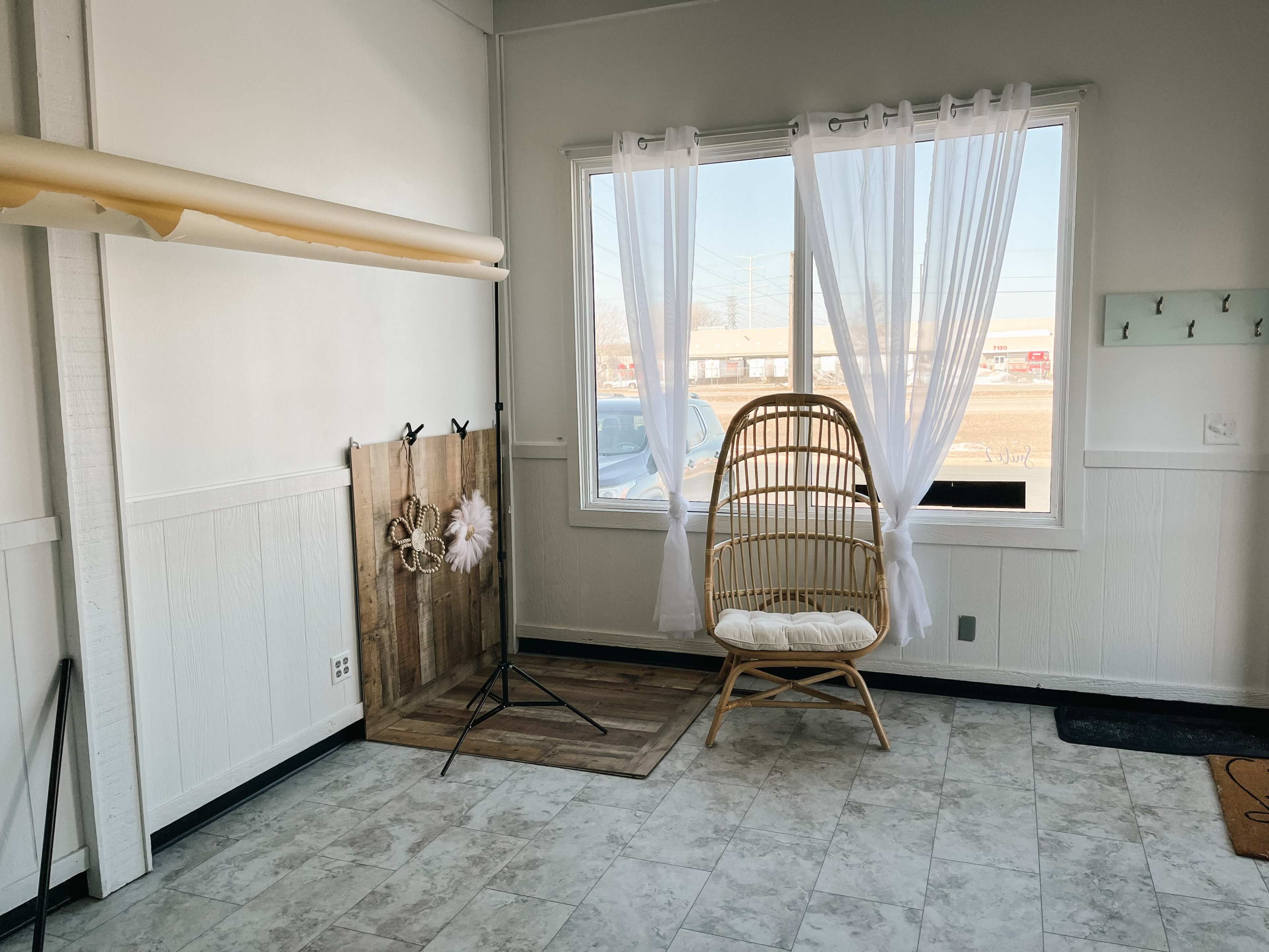 A brightly lit room features a rattan chair next to a window, with sheer curtains and a wooden backdrop.