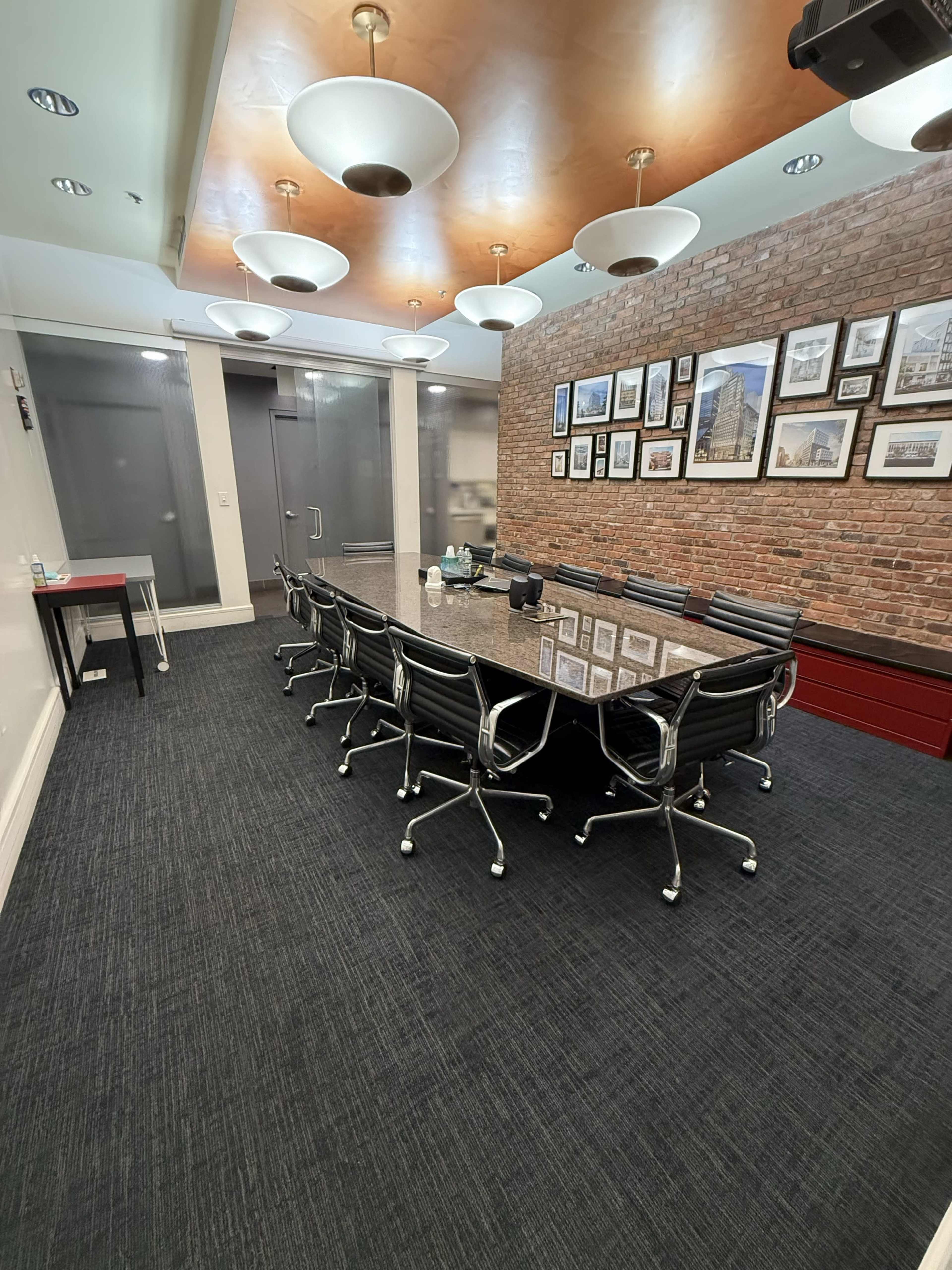 The image shows a conference room featuring a long table surrounded by black chairs, with a brick wall adorned with framed pictures and a ceiling lined with pendant lights.