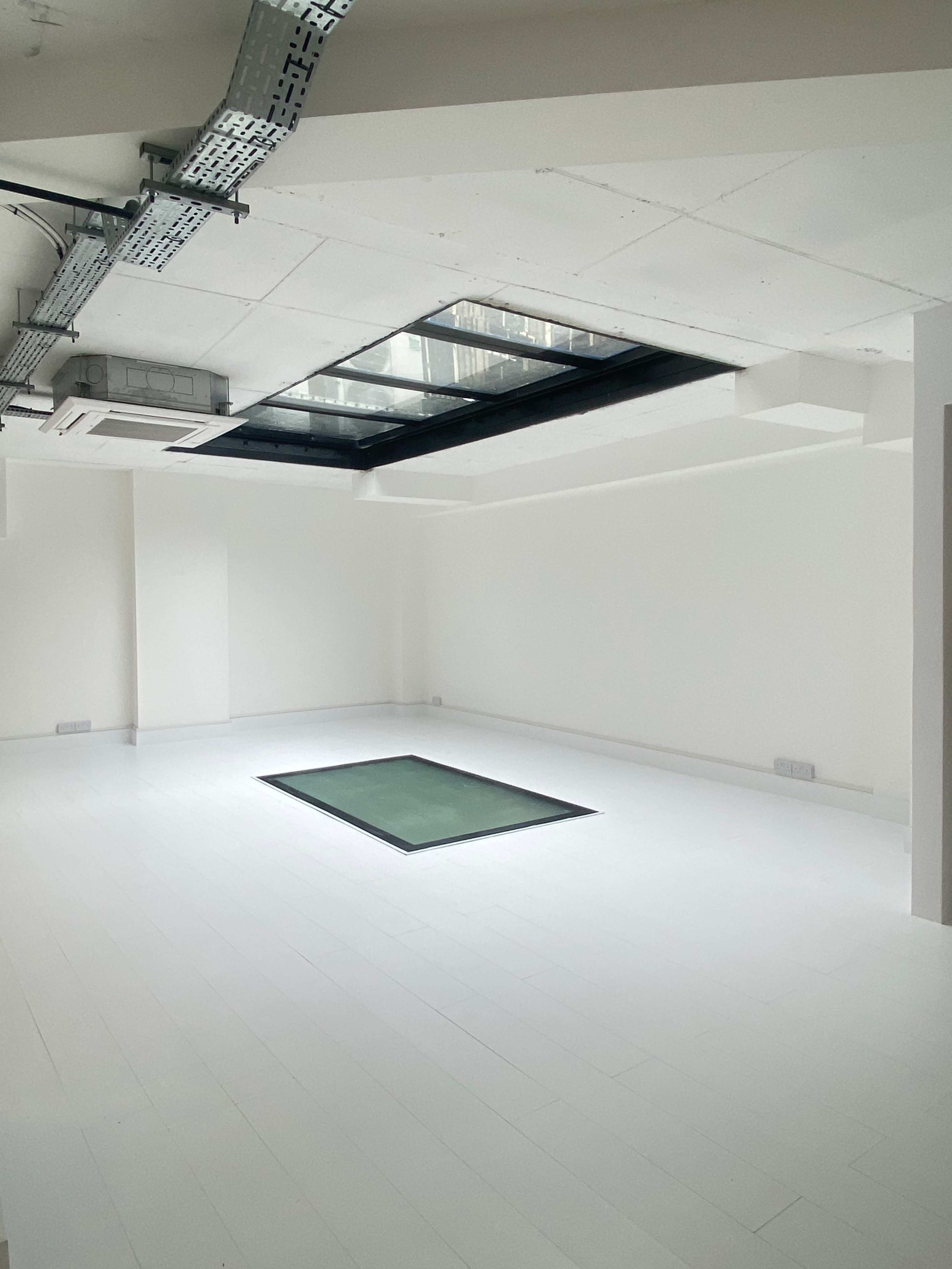 A spacious, empty room with white walls and flooring, featuring a square green mat in the center and a skylight above.