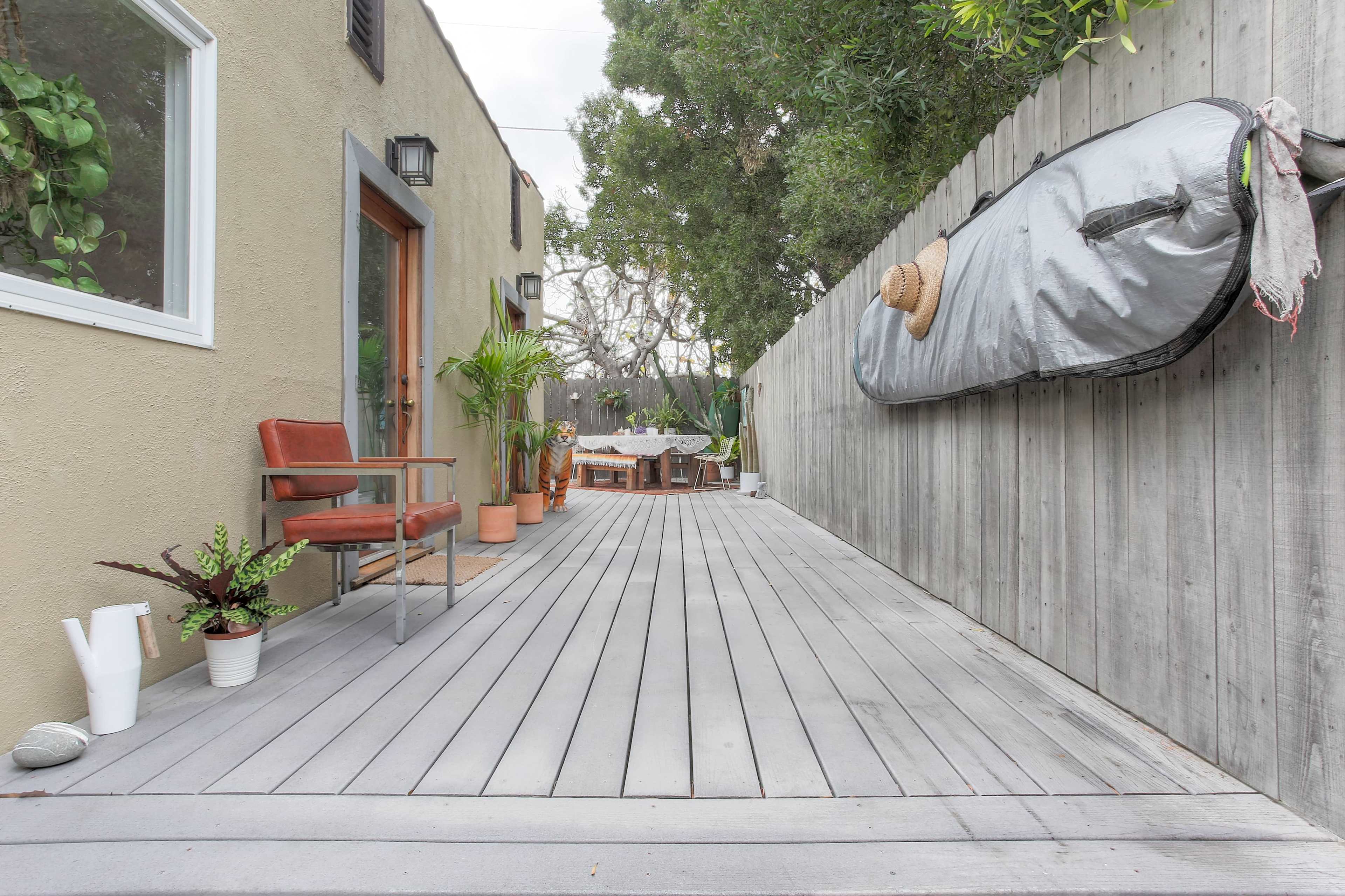 A narrow wooden deck runs alongside a house, lined with potted plants and a surfboard secured to the fence.