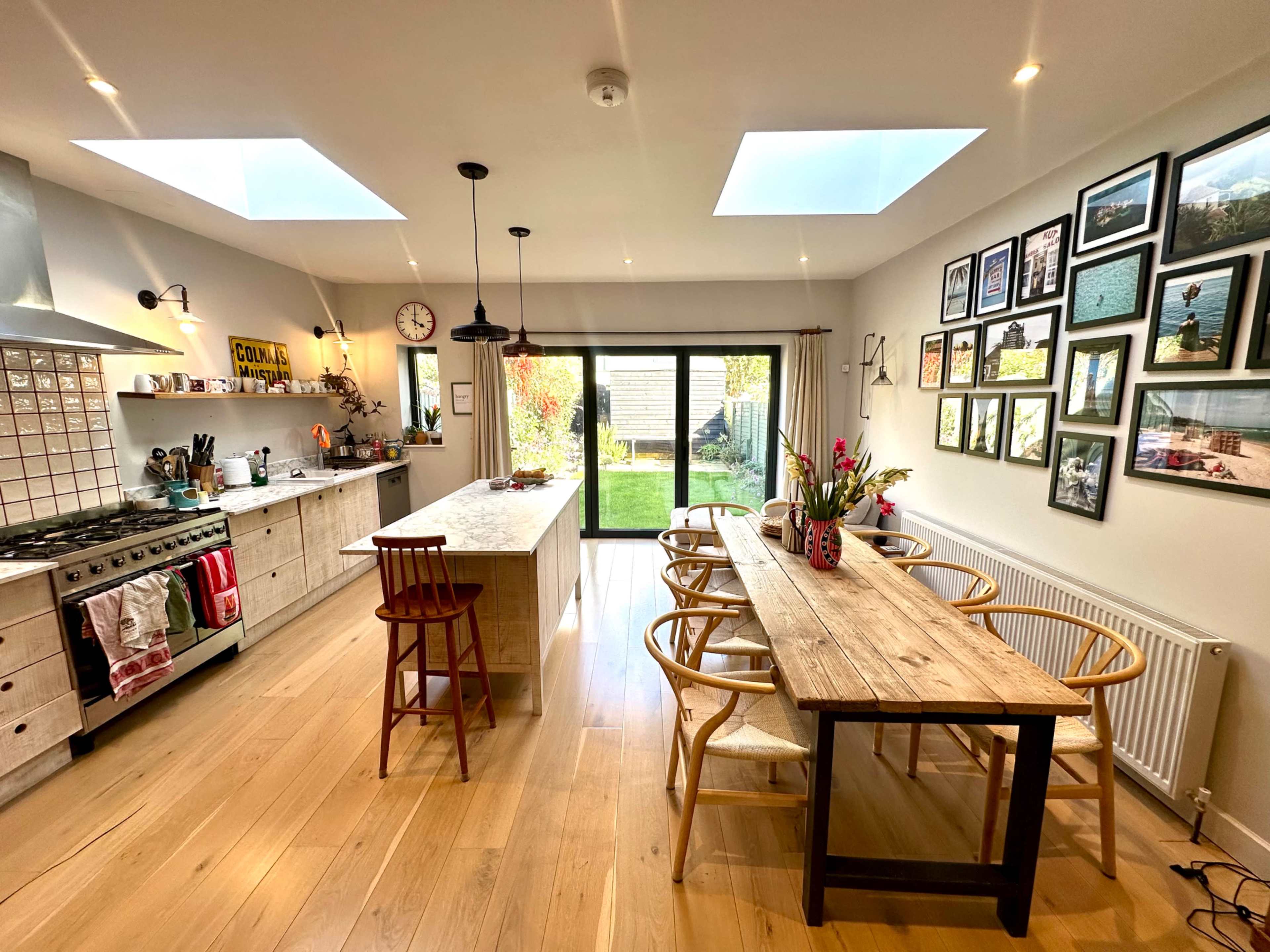 The image shows a modern kitchen and dining area with an island, a wooden dining table, and wall-mounted framed photos, illuminated by natural light through skylights and large glass doors leading to a garden.
