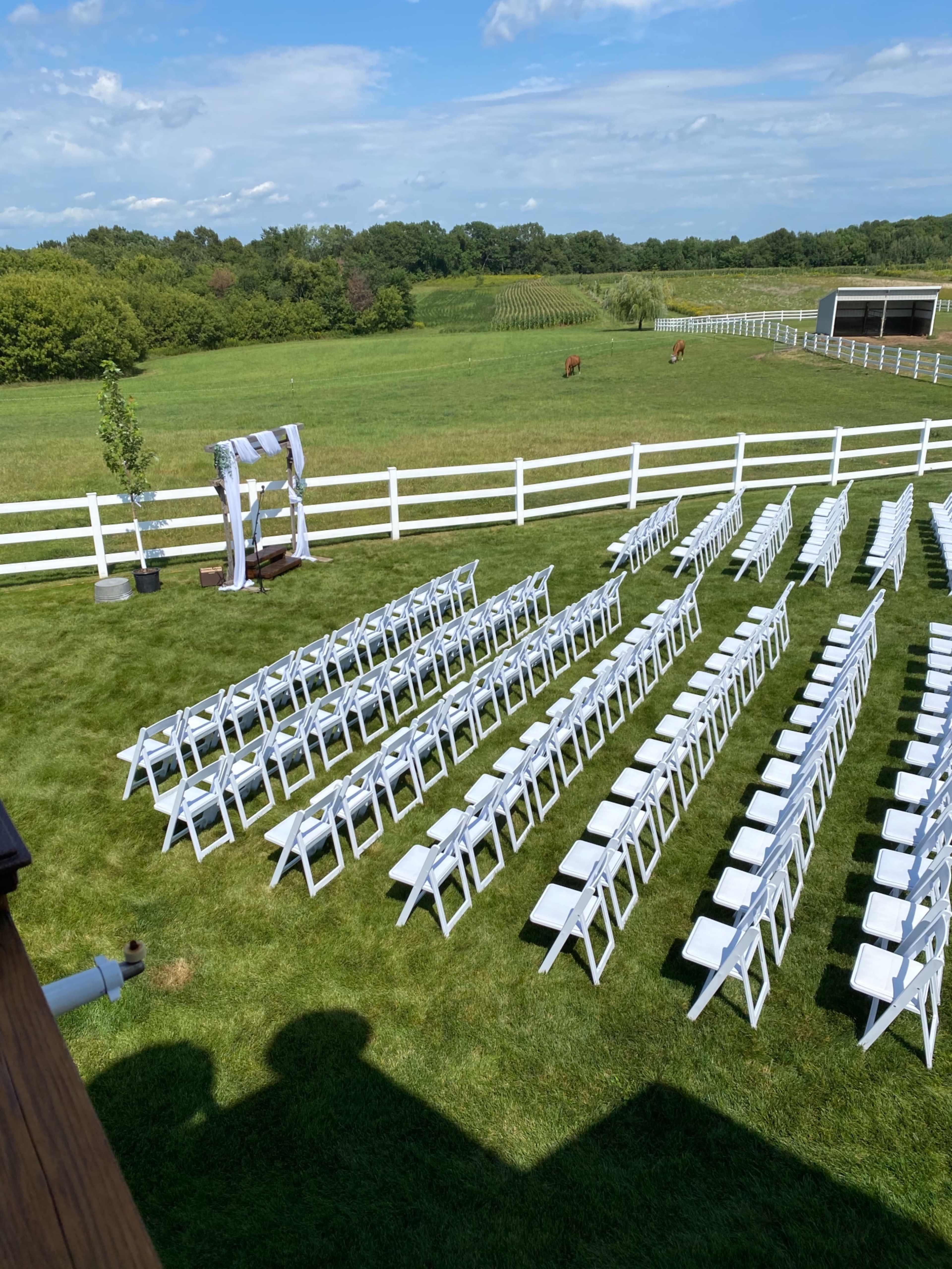 A neatly arranged outdoor wedding setup with white chairs facing an archway on a lawn surrounded by greenery and a white fence.