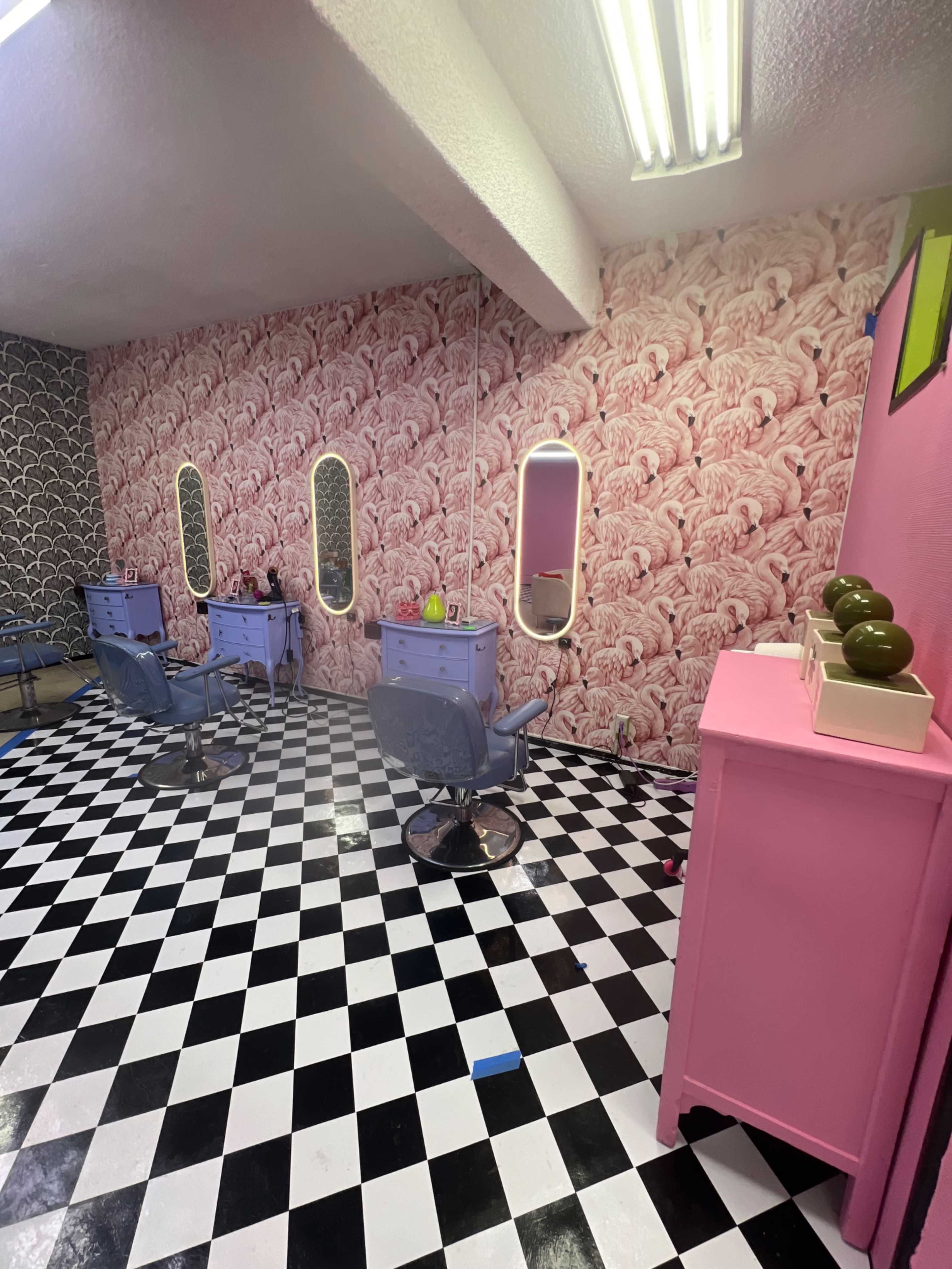 A modern hair salon with pink and black decor, featuring multiple styling stations with mirrors, blue chairs, and a checkered floor.
