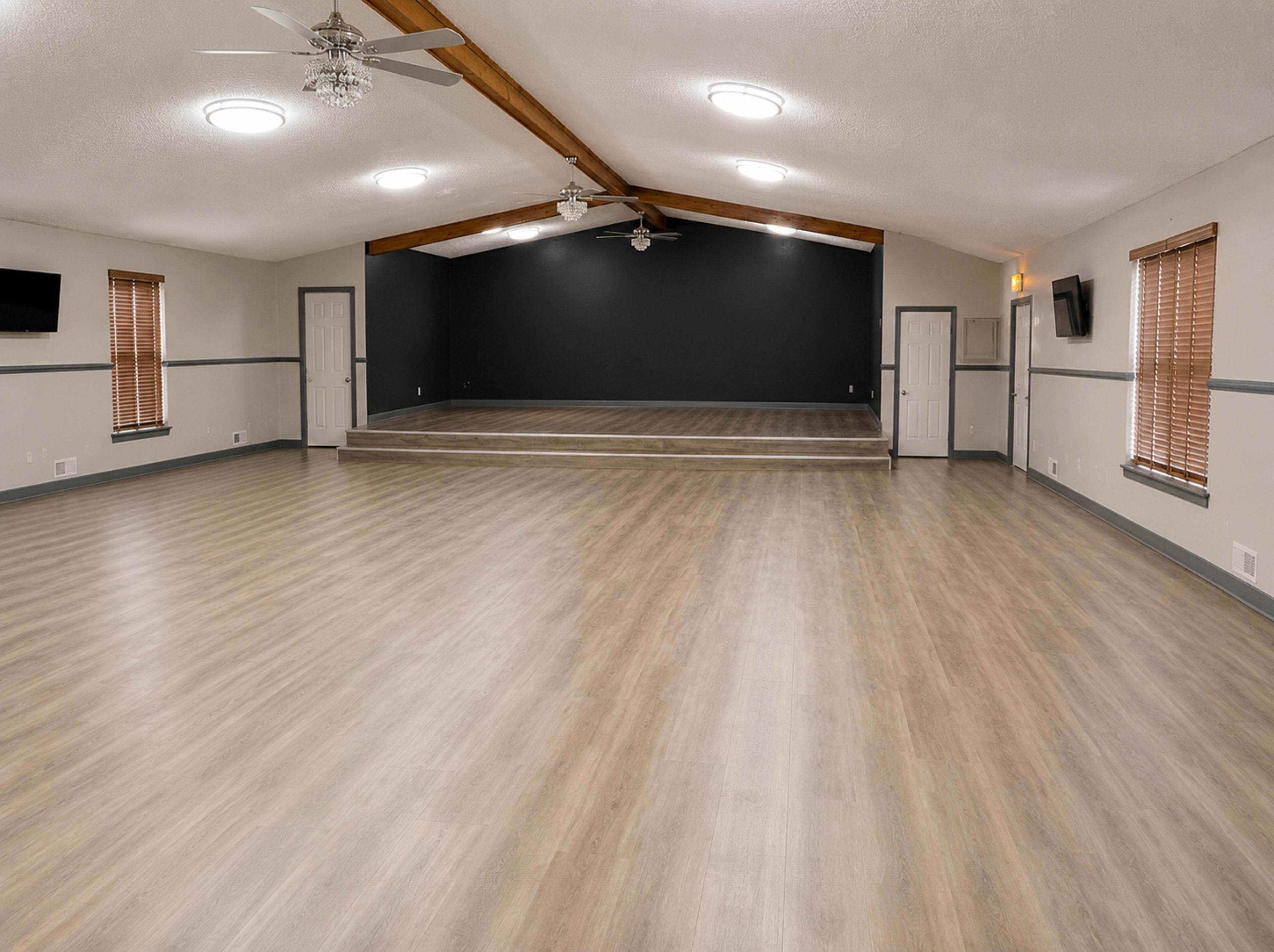 The image shows a spacious, empty room with wooden flooring, a high ceiling supported by beams, and a raised stage against a dark wall.