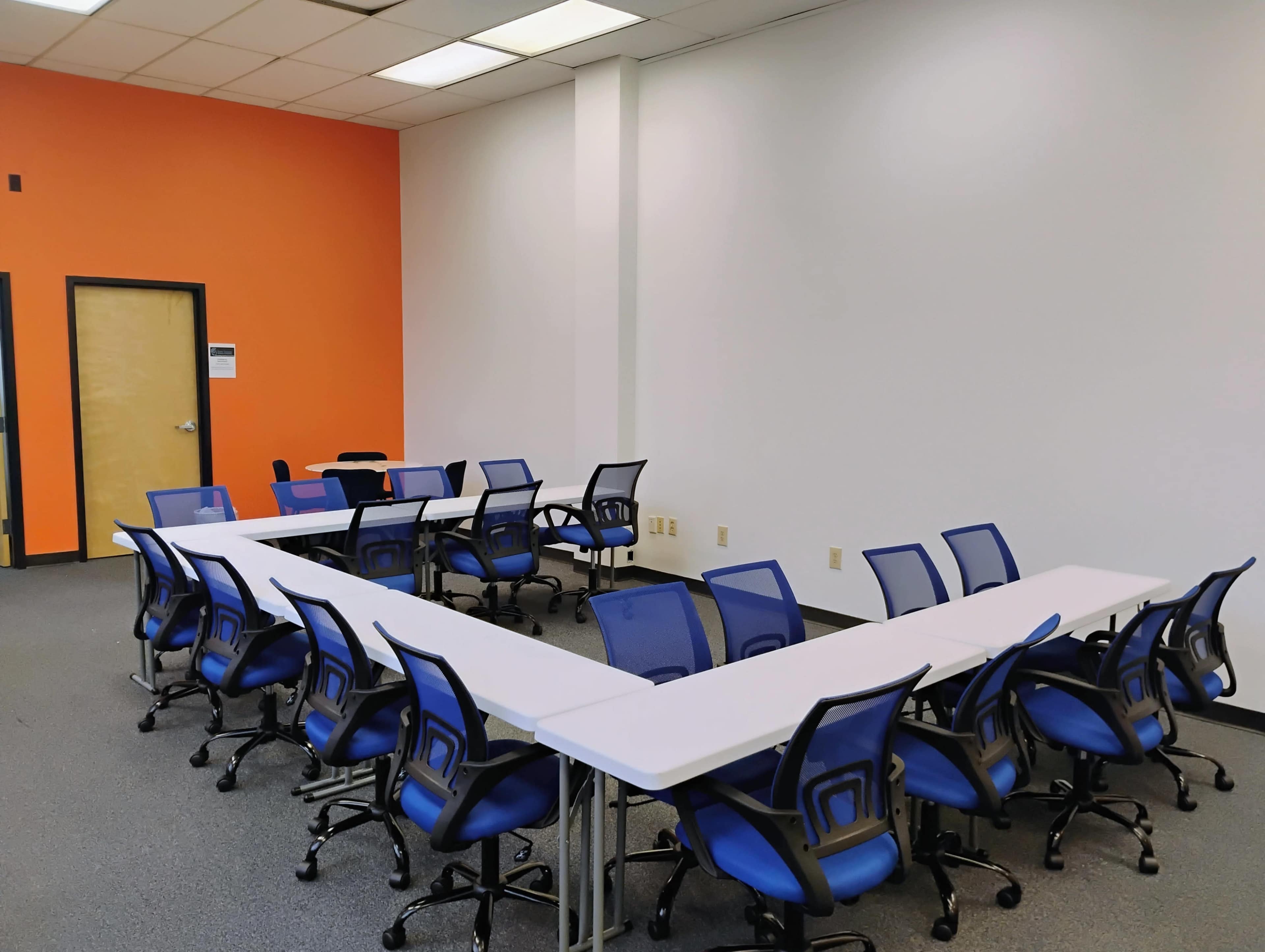 A conference room features multiple blue rolling chairs arranged around white tables in a U-shape against an orange wall.
