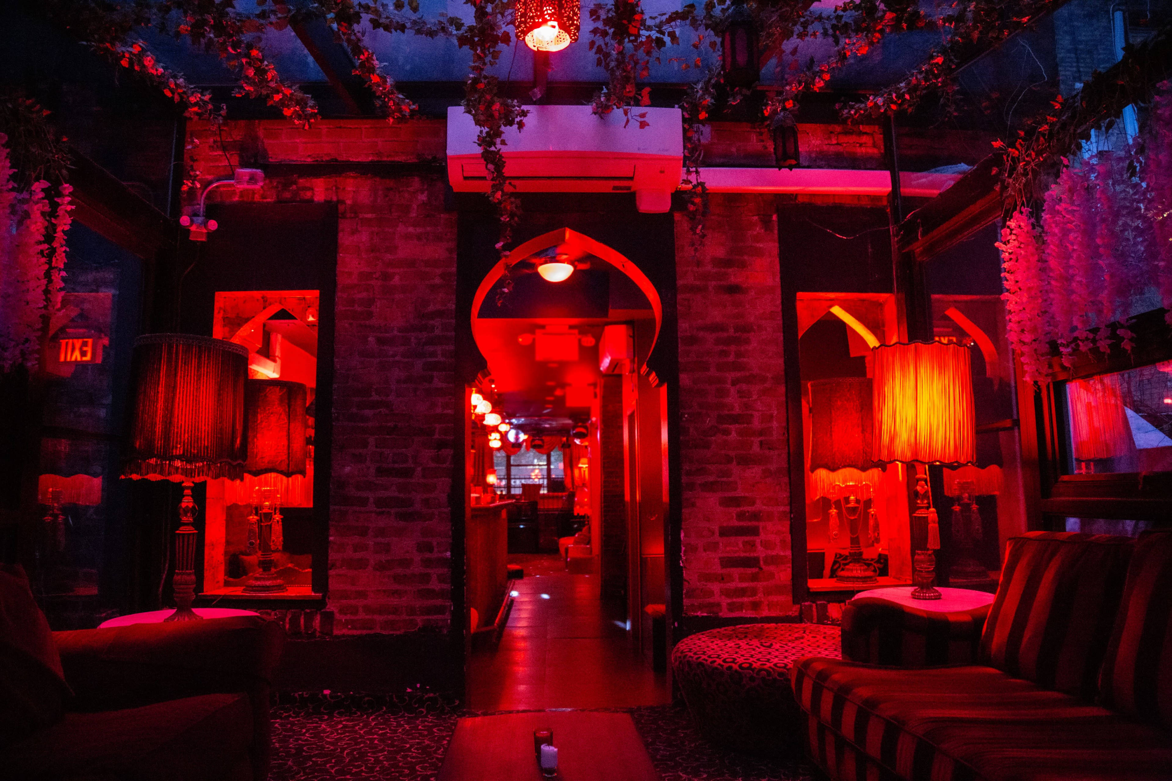 An intimate lounge with red lighting, decorated with vintage lamps and lush greenery, leading to a bar area.