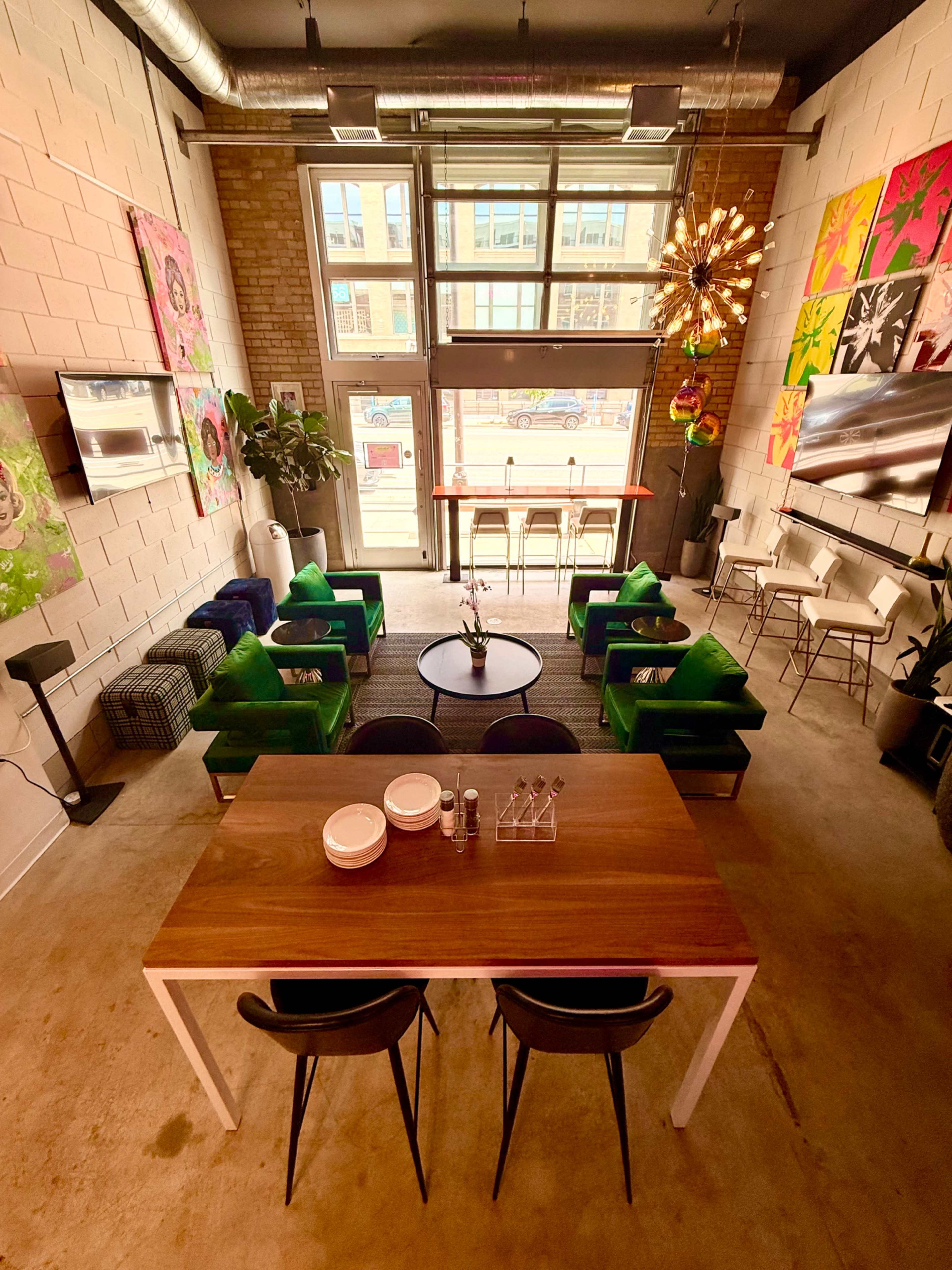 The image shows a modern café interior with green armchairs, a wooden dining table, and large windows providing natural light.
