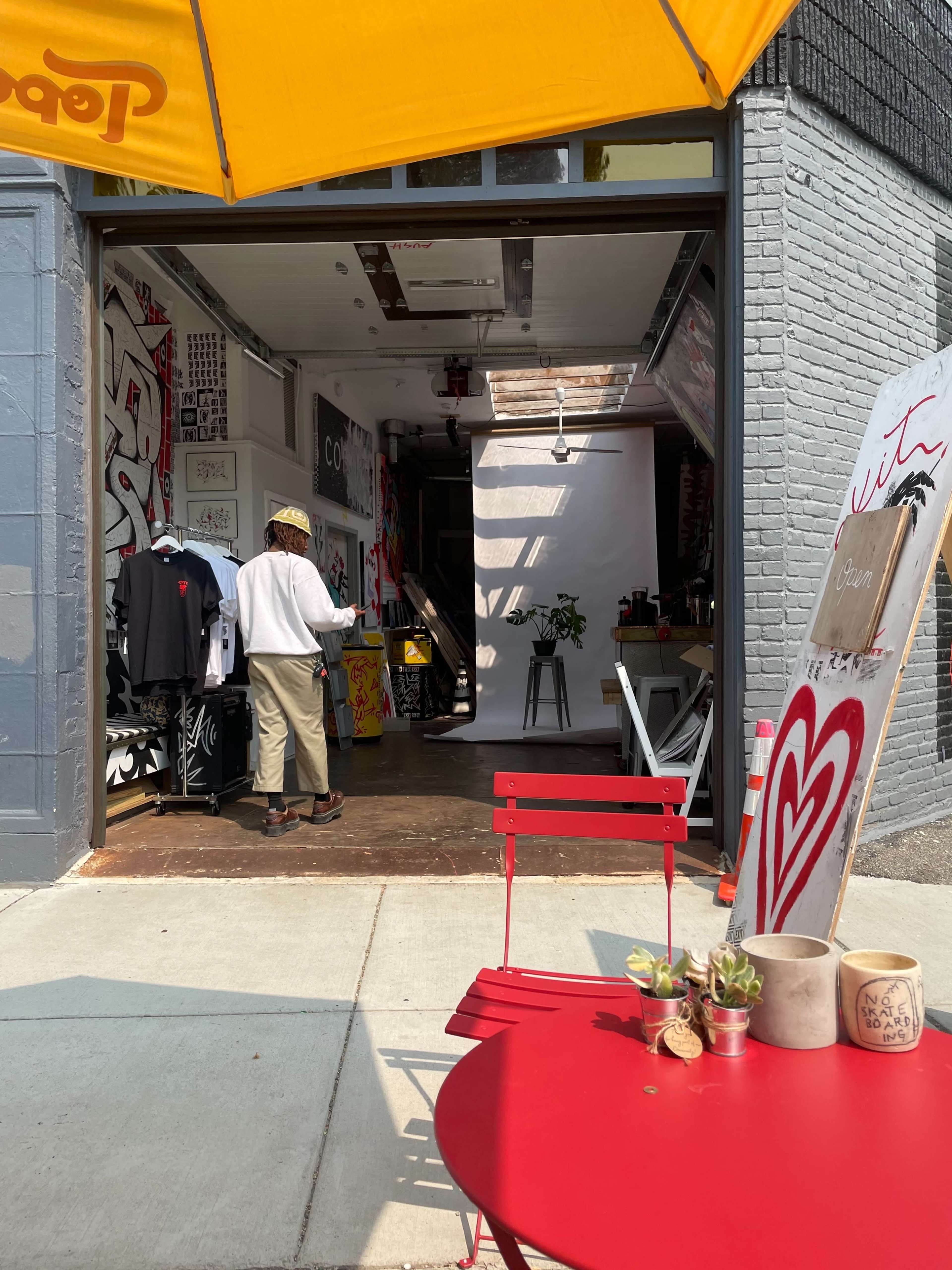 A person in a straw hat stands inside a storefront filled with art and clothing, while a red table and chairs are placed on the sidewalk outside.
