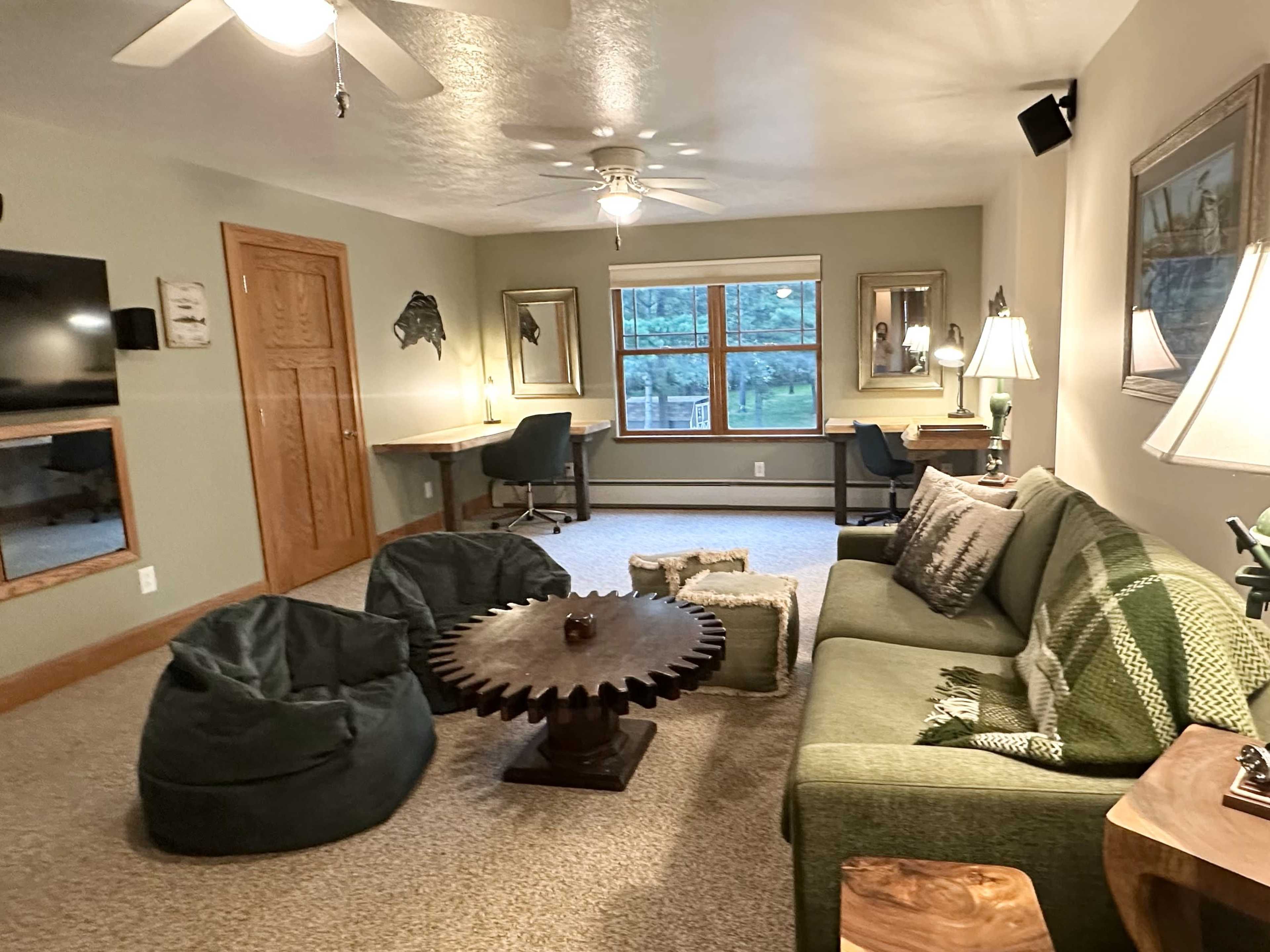 The room features a green sofa, bean bag chairs, a round wooden coffee table, and a desk by a window, all arranged in a cozy, well-lit setting.