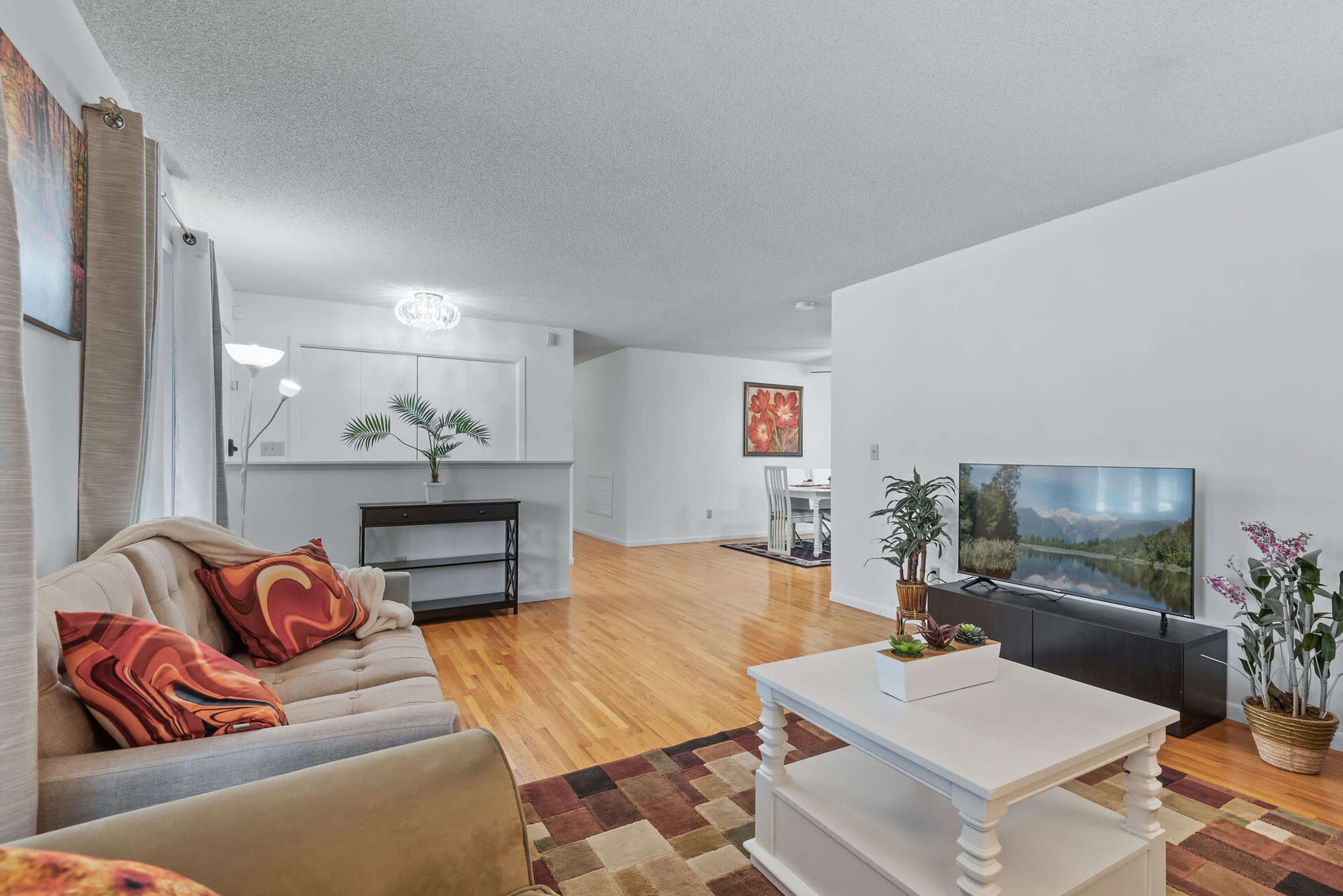 A modern living room features a beige sofa with colorful cushions, a white coffee table, a television on a stand, and wooden flooring.