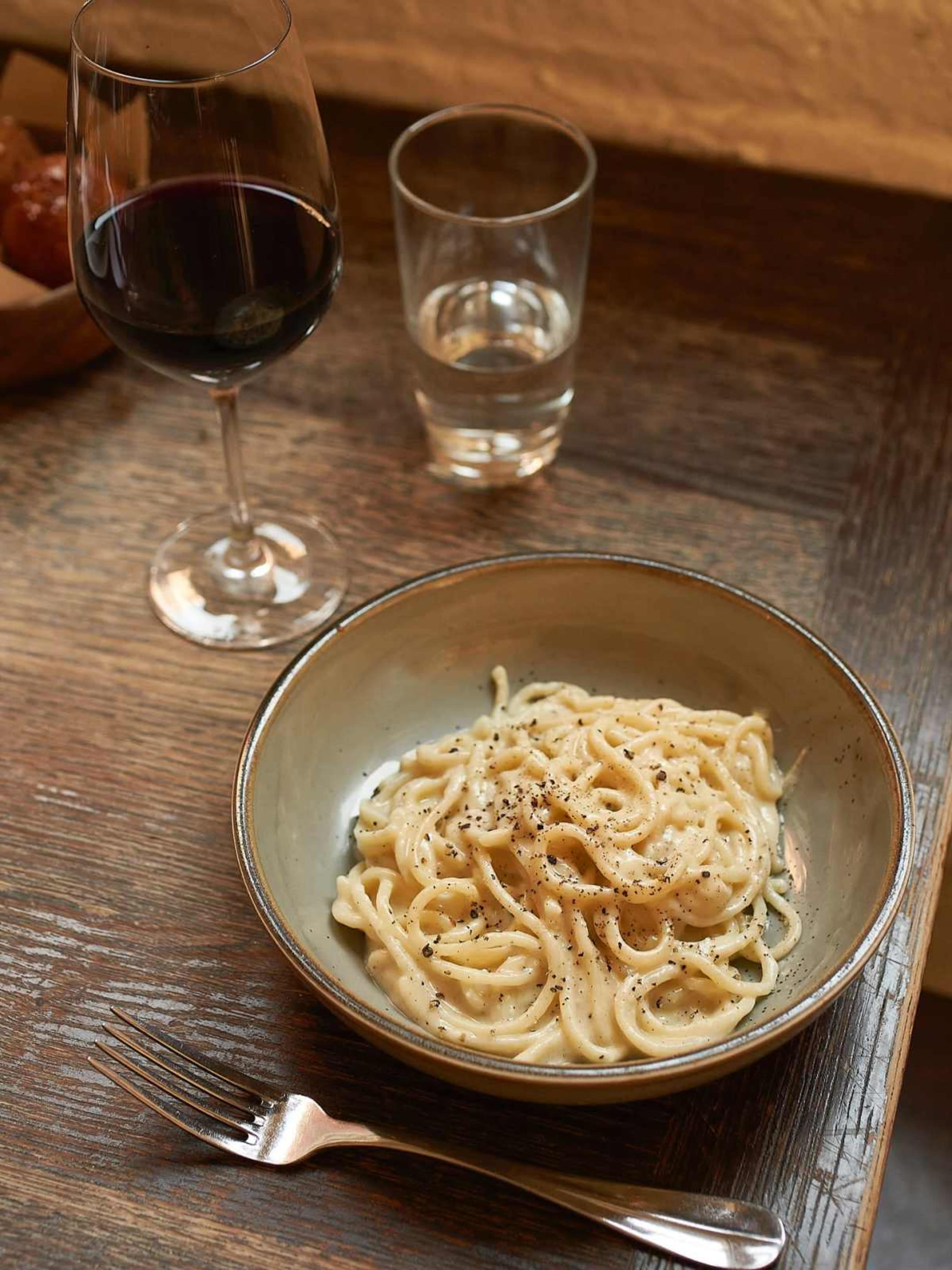 A bowl of spaghetti with creamy sauce and black pepper is placed on a wooden table next to a glass of red wine and a glass of water.