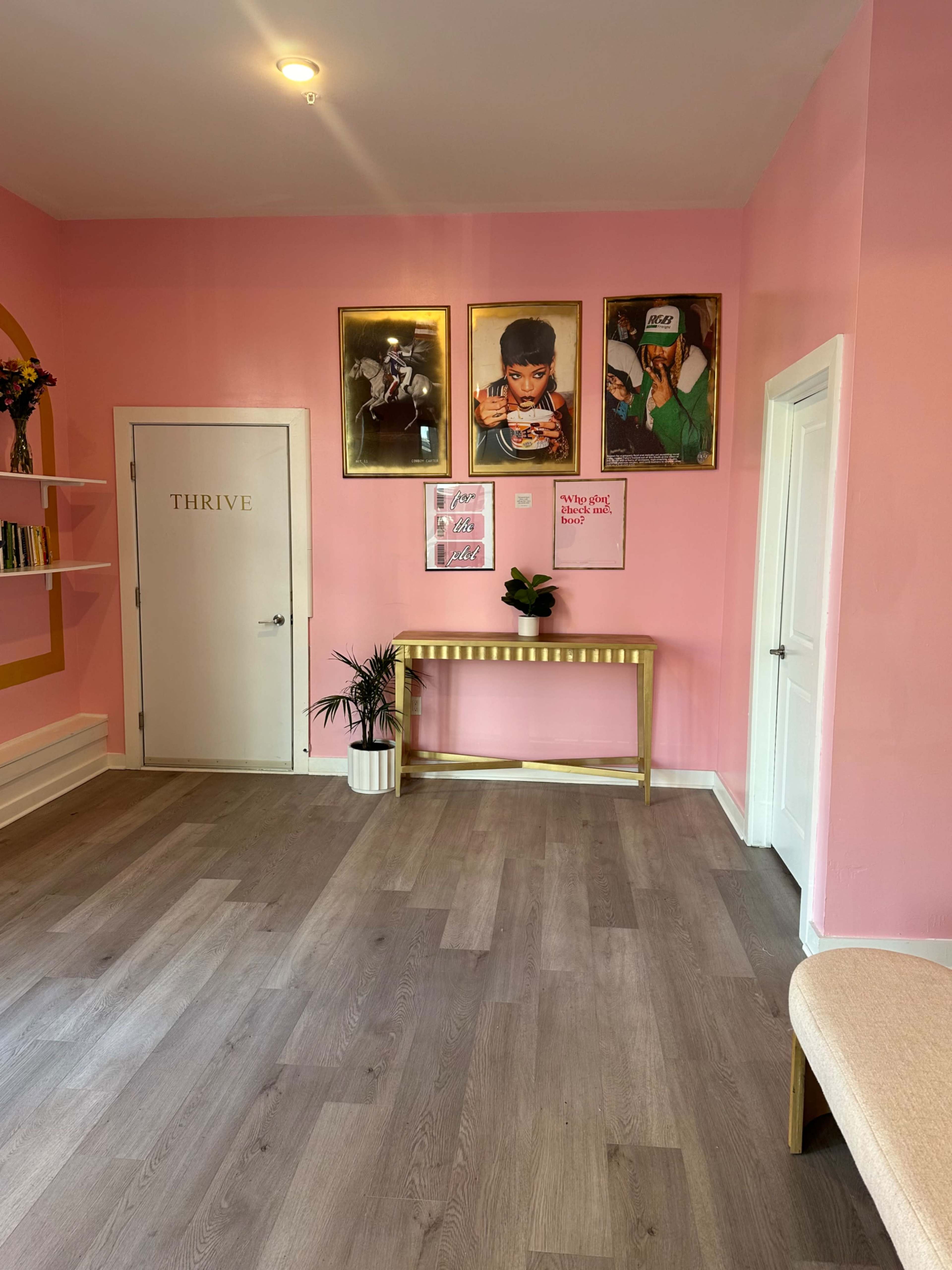 A pink-walled room featuring framed artwork, a gold console table, a plant, and a door labeled "THRIVE."