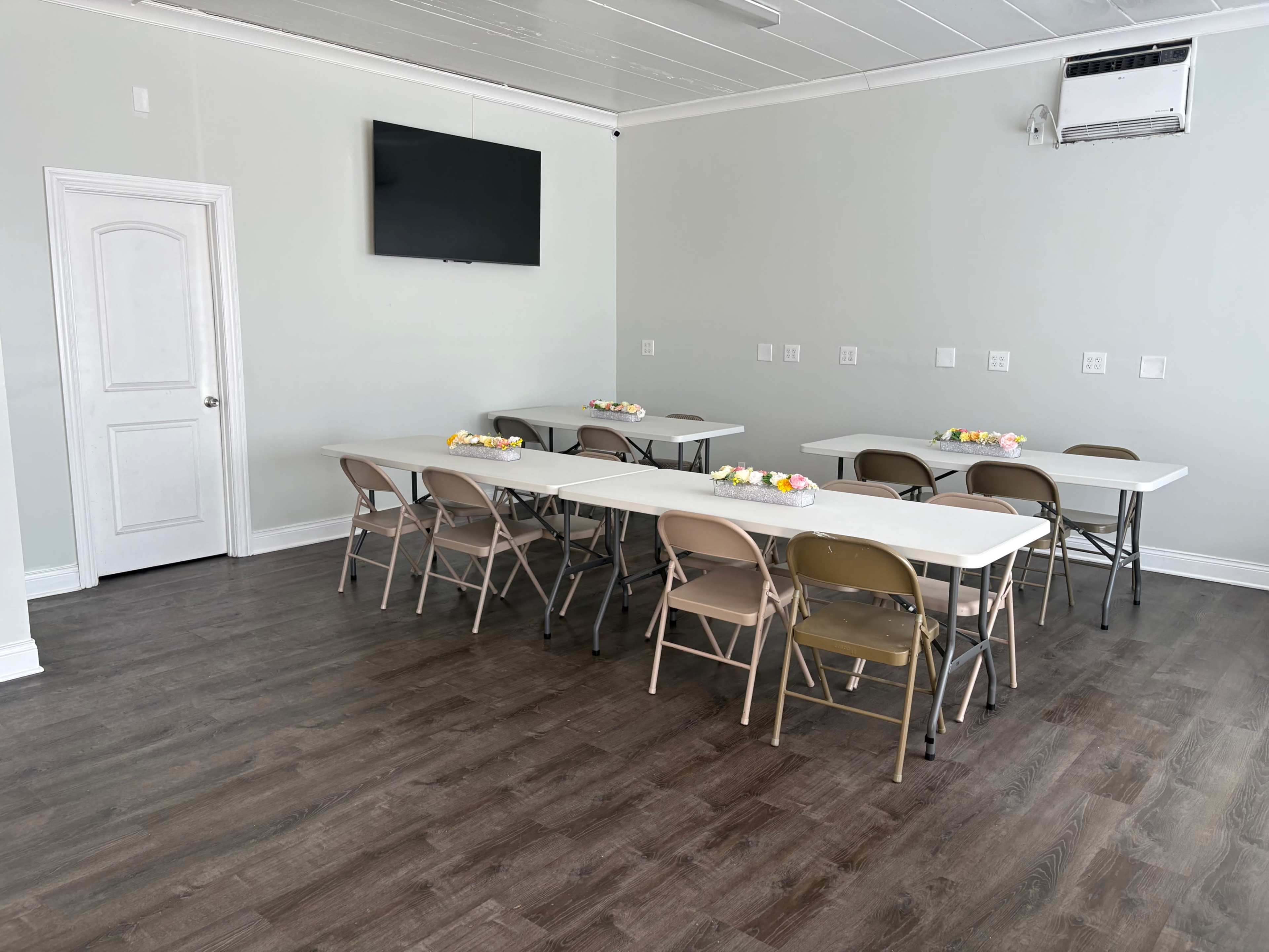 The image shows a light-colored room with gray walls, featuring several folding tables arranged with chairs and a mounted television on one wall.