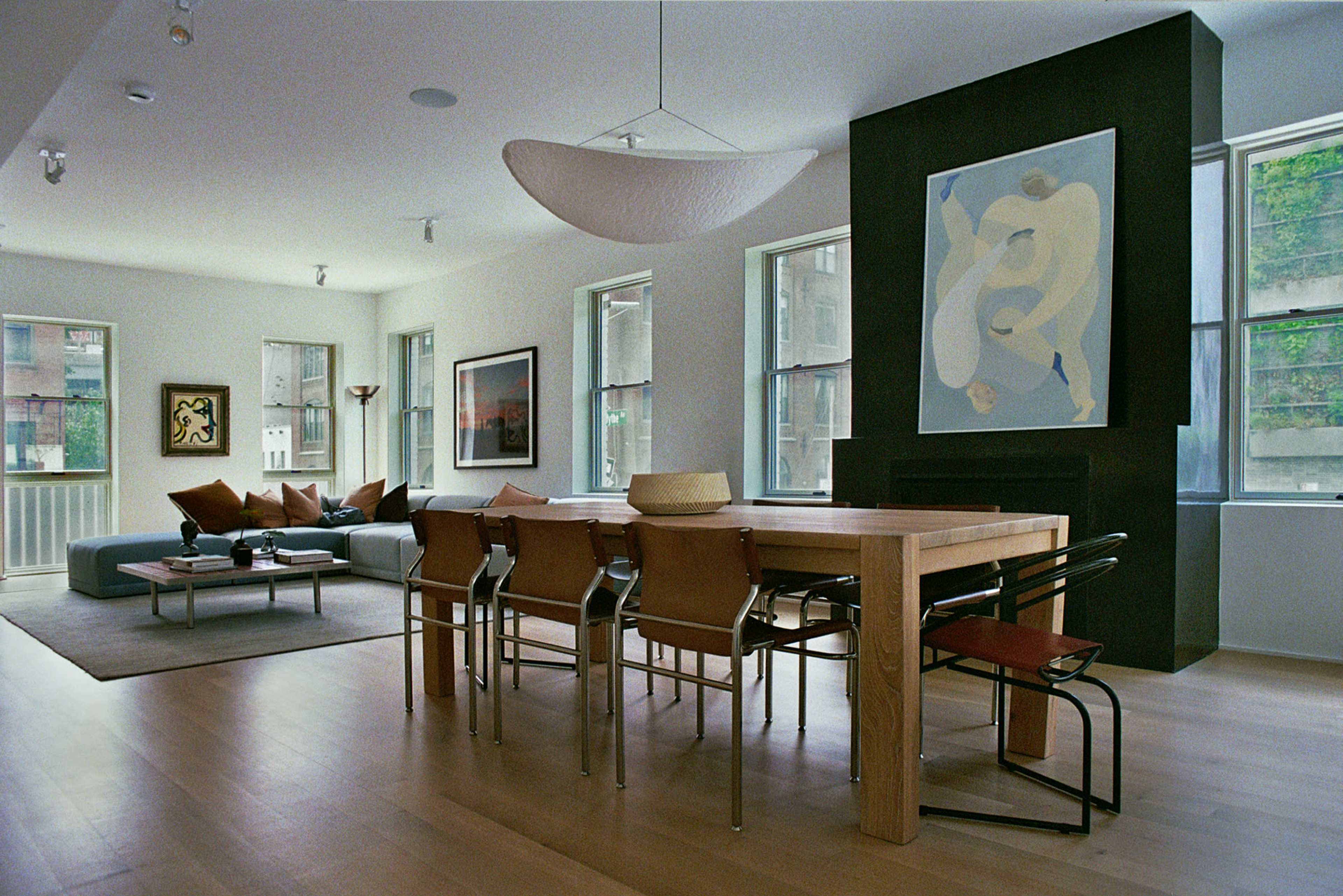 The image shows a modern, open living space featuring a dining area with a large wooden table and several chairs, alongside a seating area with a sofa, artworks on the walls, and large windows allowing natural light.