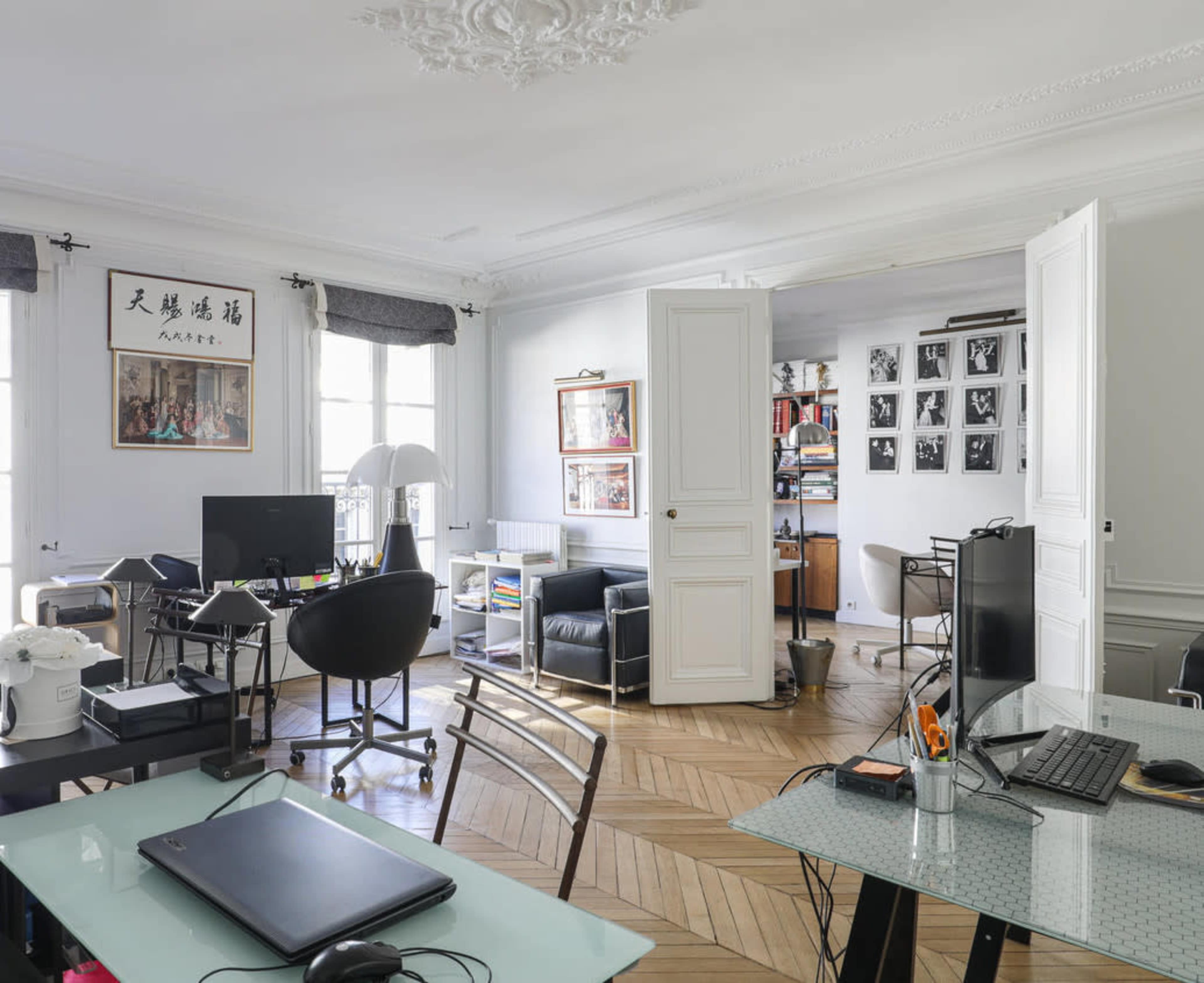 A modern office space features multiple workstations, a variety of chairs, and a separation between two rooms, with art displayed on the walls.