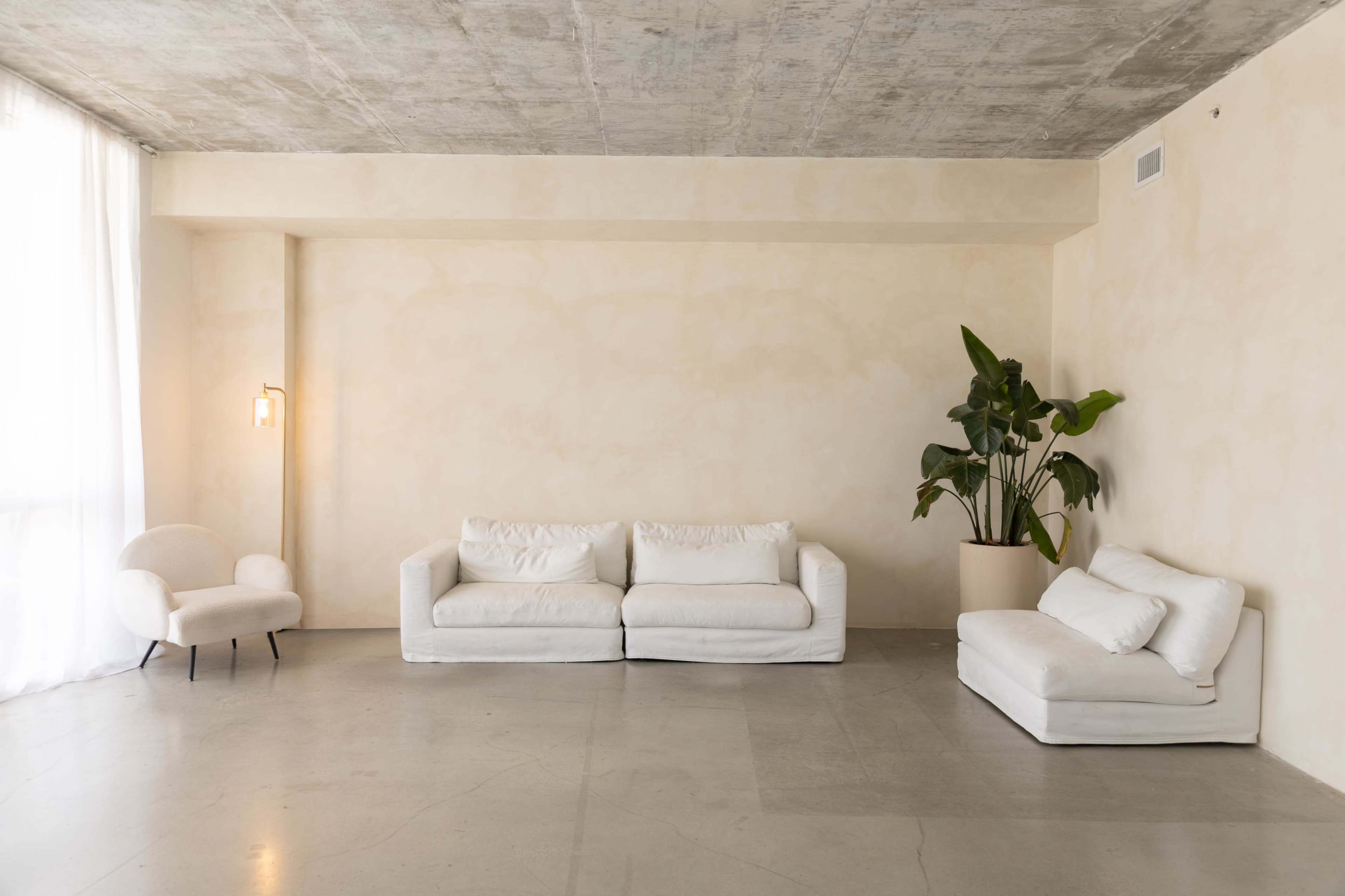 The image shows a minimalist living room with two white sofas, a small armchair, and a large green plant in a beige pot against a light, textured wall.