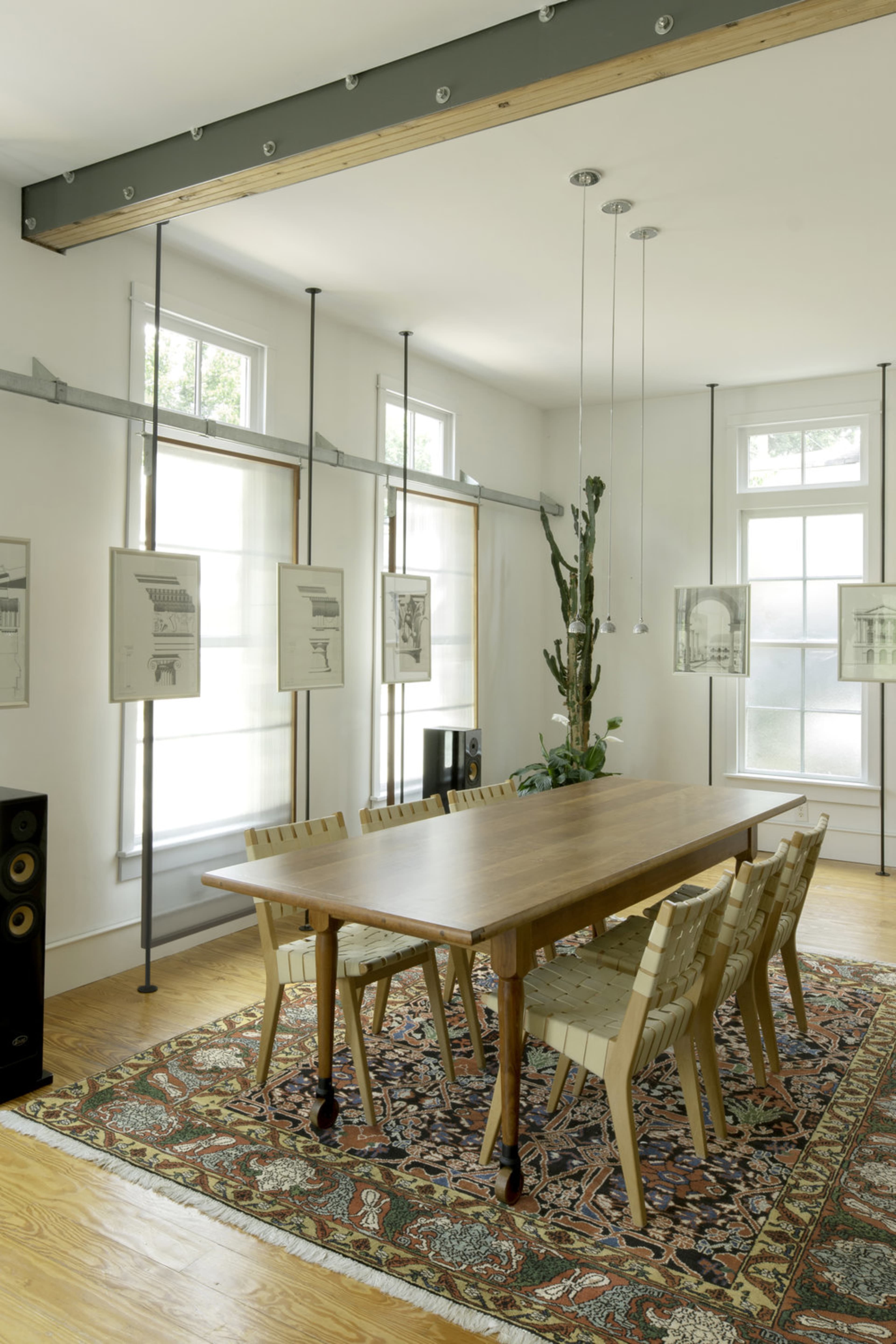 A wooden dining table surrounded by six chairs sits on a patterned rug in a well-lit room with large windows and hanging artwork.