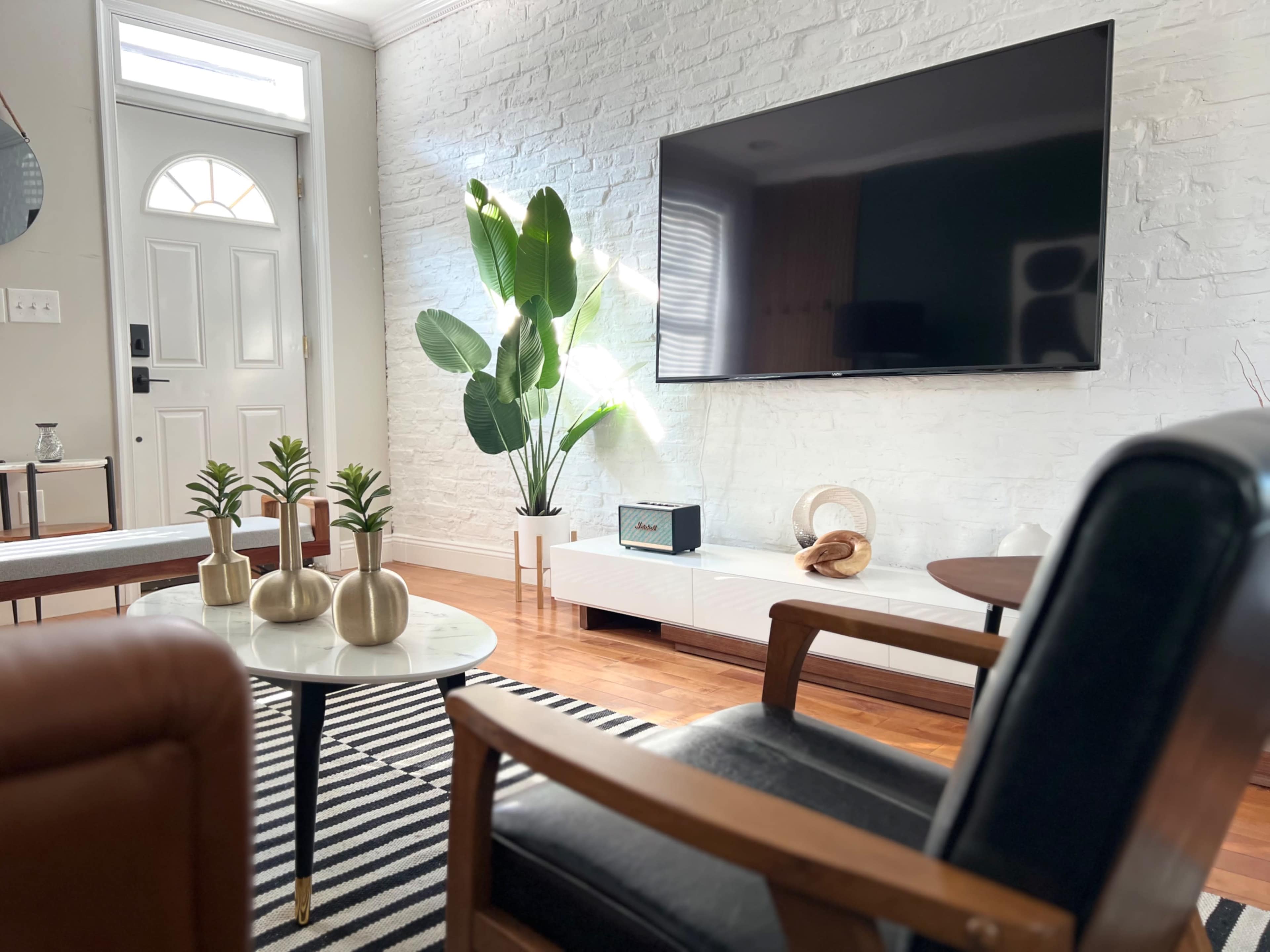 A living room features a large wall-mounted television, a coffee table with decorative plants, and a leather chair, all arranged around a striped rug.