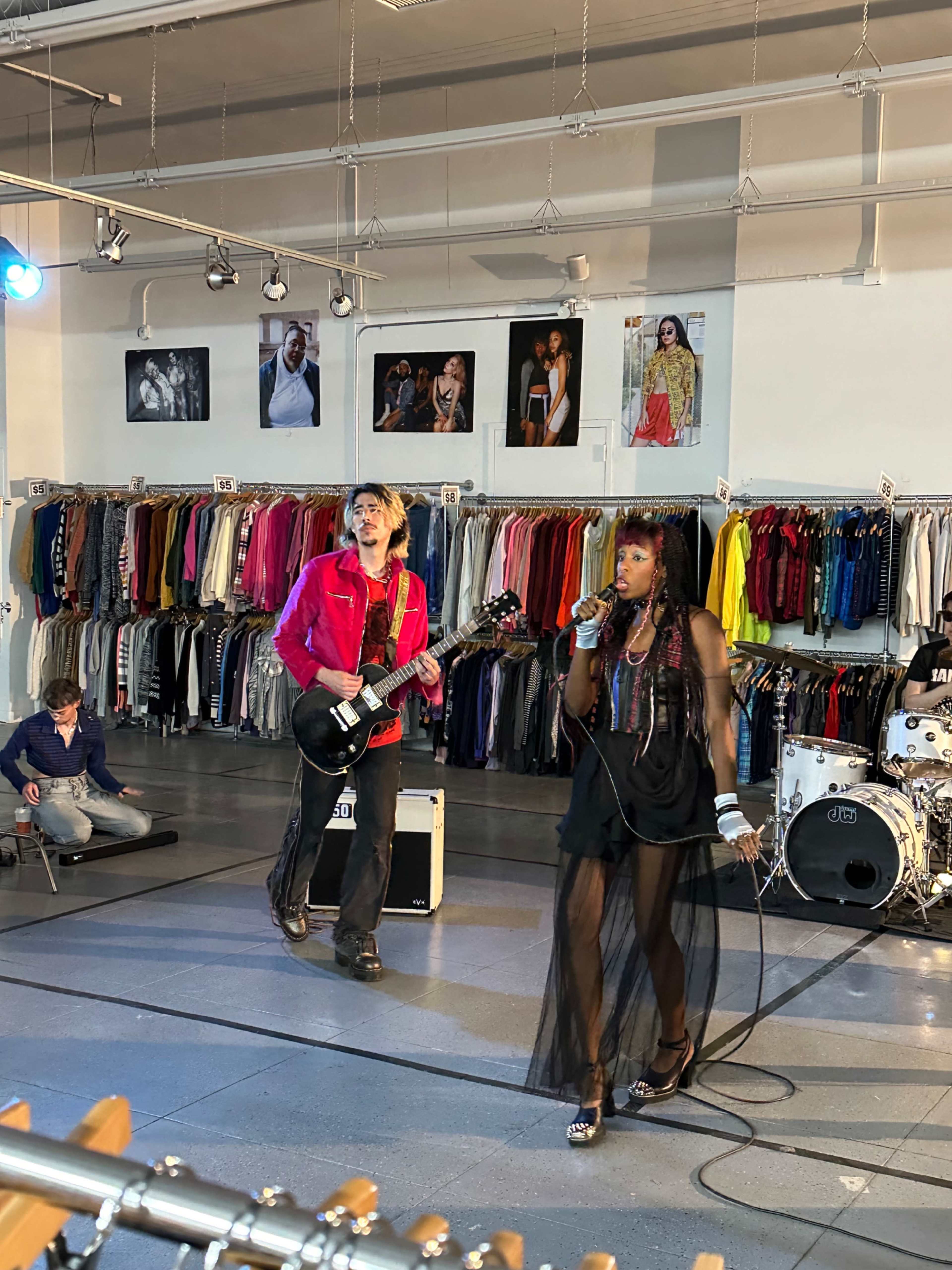 A man playing an electric guitar and a woman singing into a microphone perform on a small stage in a clothing store filled with hanging garments.
