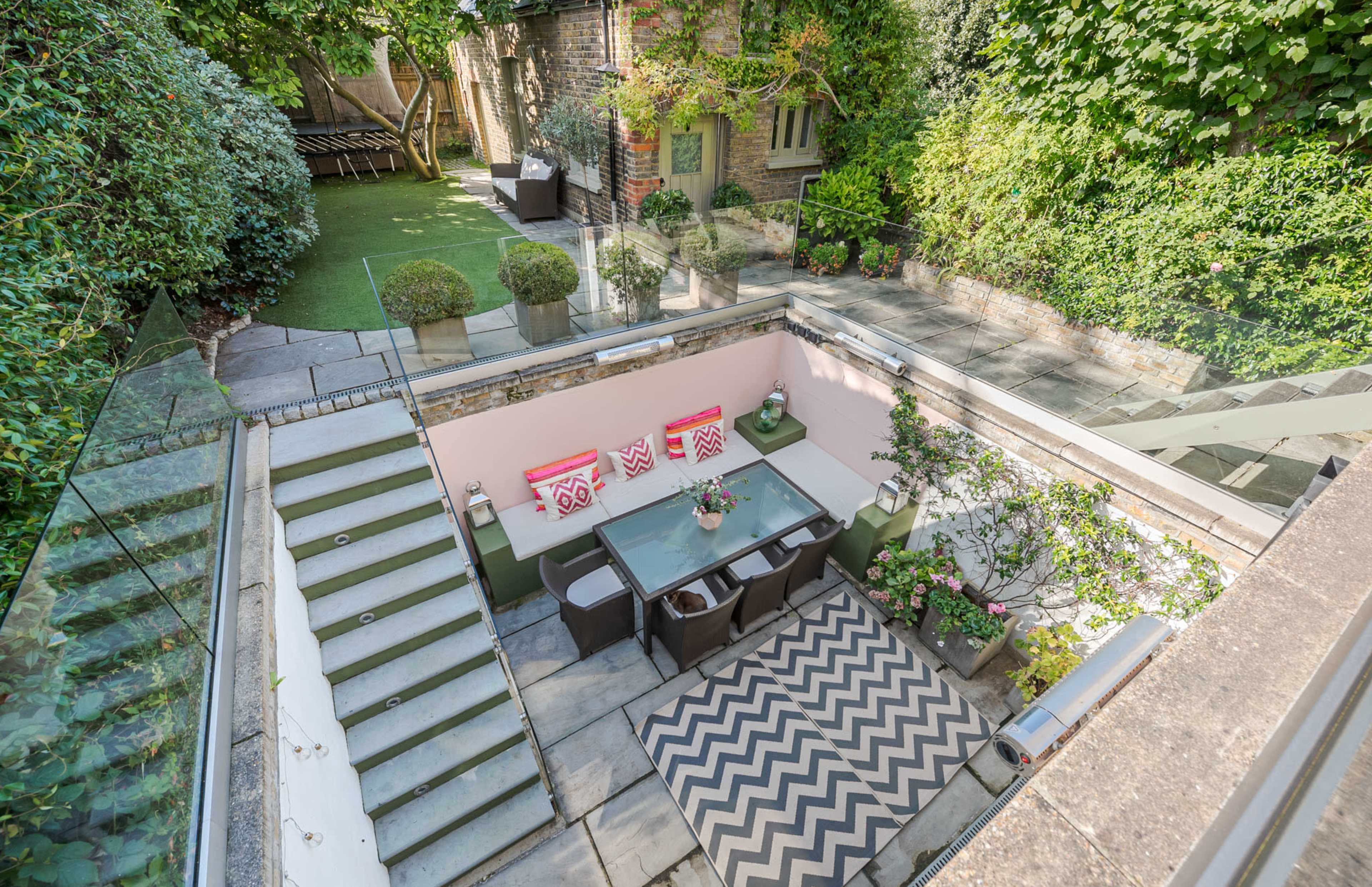 A sunlit outdoor dining area with a glass table surrounded by chairs, set on a patterned rug, and framed by lush greenery and a staircase leading to a garden.