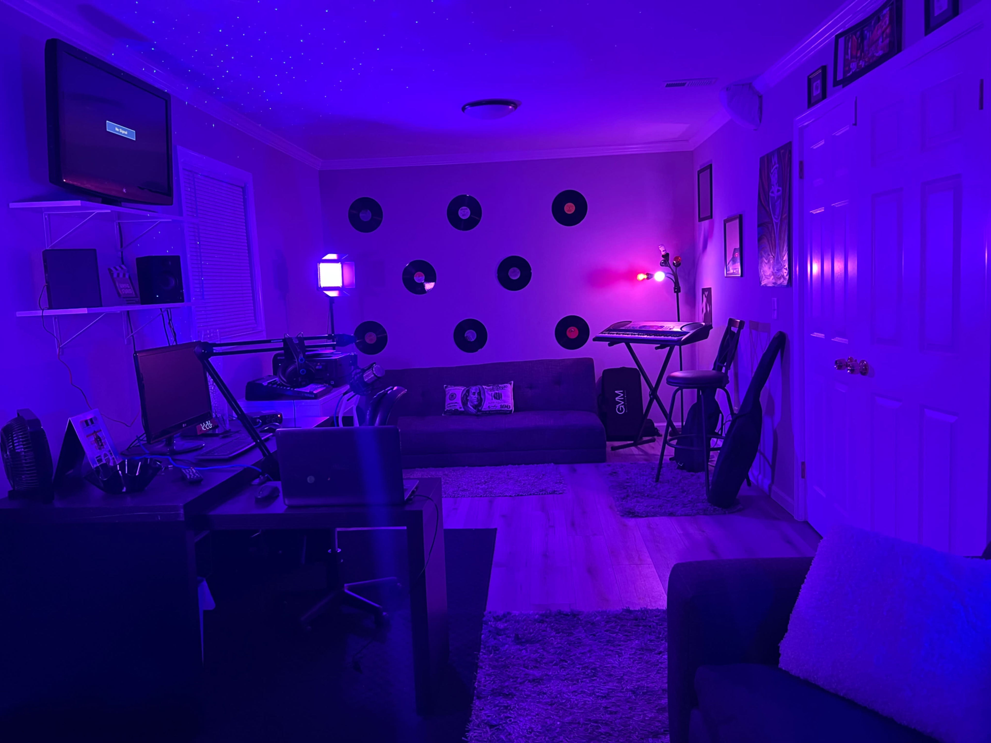 The image shows a dimly lit room with purple lighting, featuring a couch, a desk with a computer and monitor, a keyboard on a stand, and vinyl records arranged on the wall.