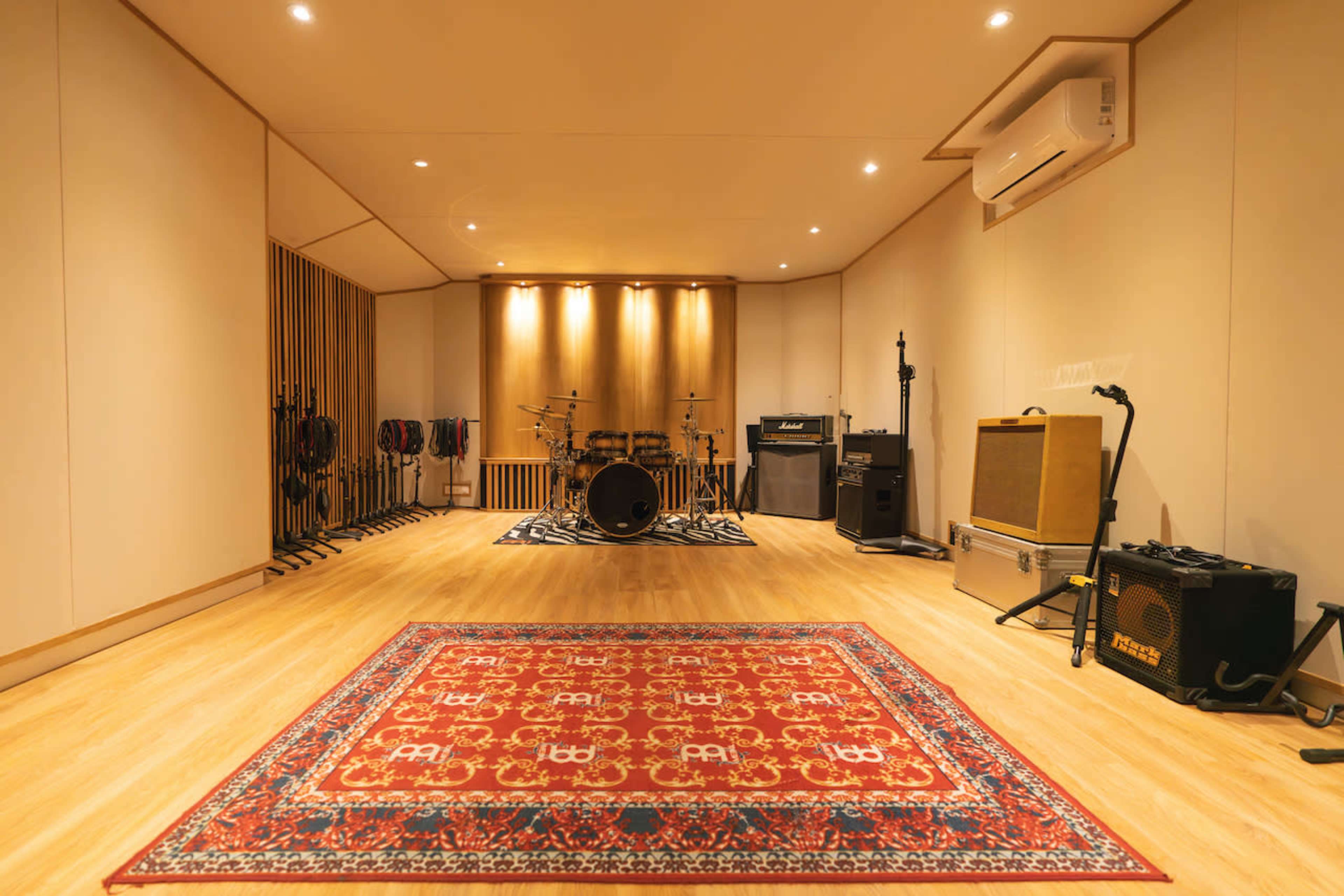 The image shows a music rehearsal studio with a drum set, amplifiers, and a prominent area rug on the wooden floor.