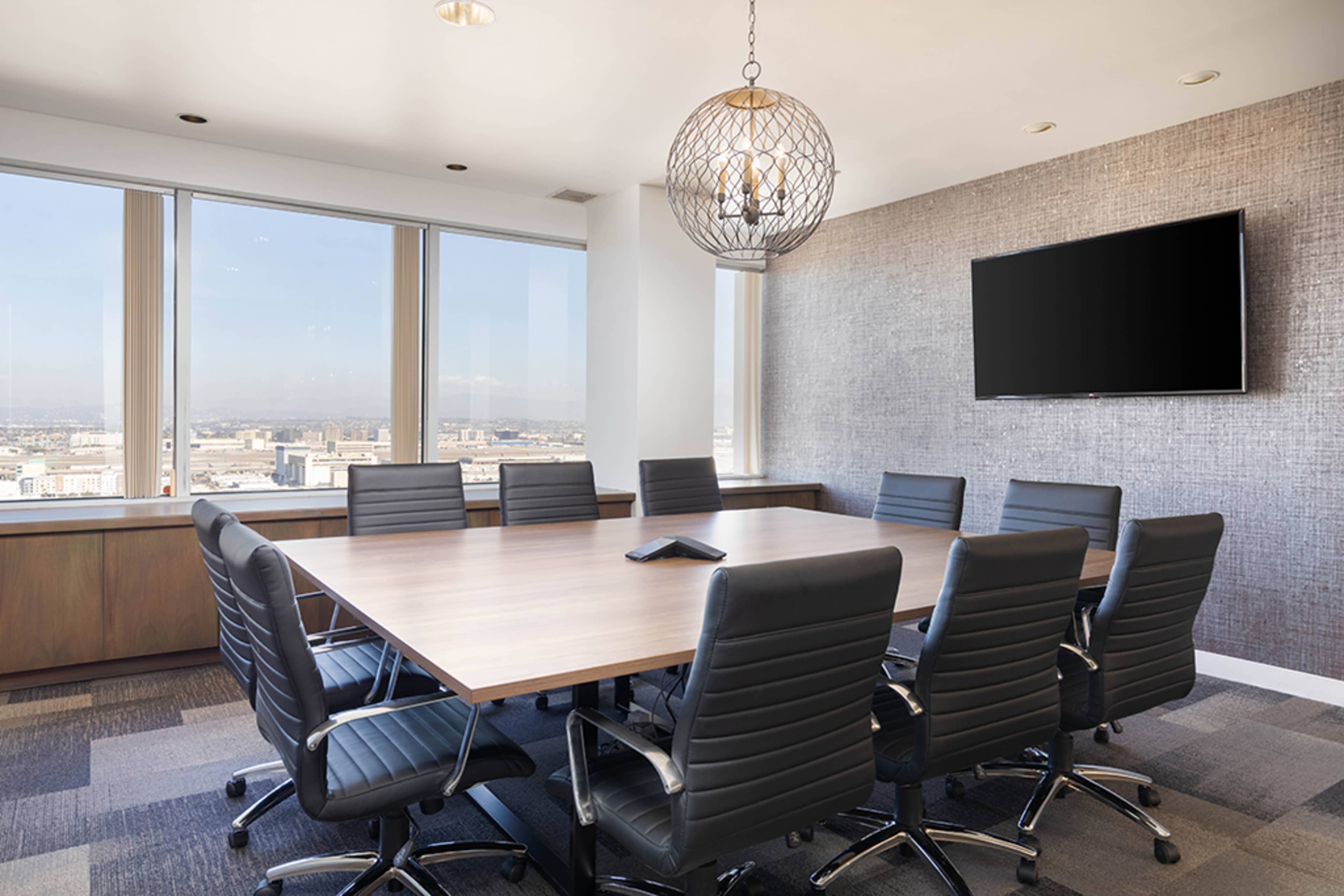 A modern conference room features a large wooden table surrounded by leather office chairs, with windows offering a view of the city and a wall-mounted television.