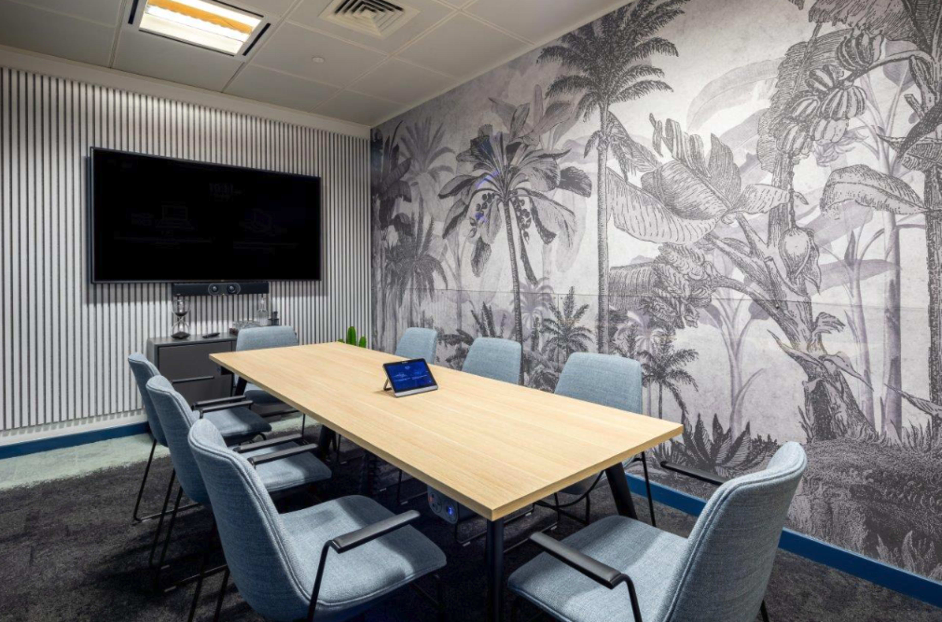 A modern conference room features a wooden table surrounded by six chairs, with a large screen and a mural of tropical plants on the wall.
