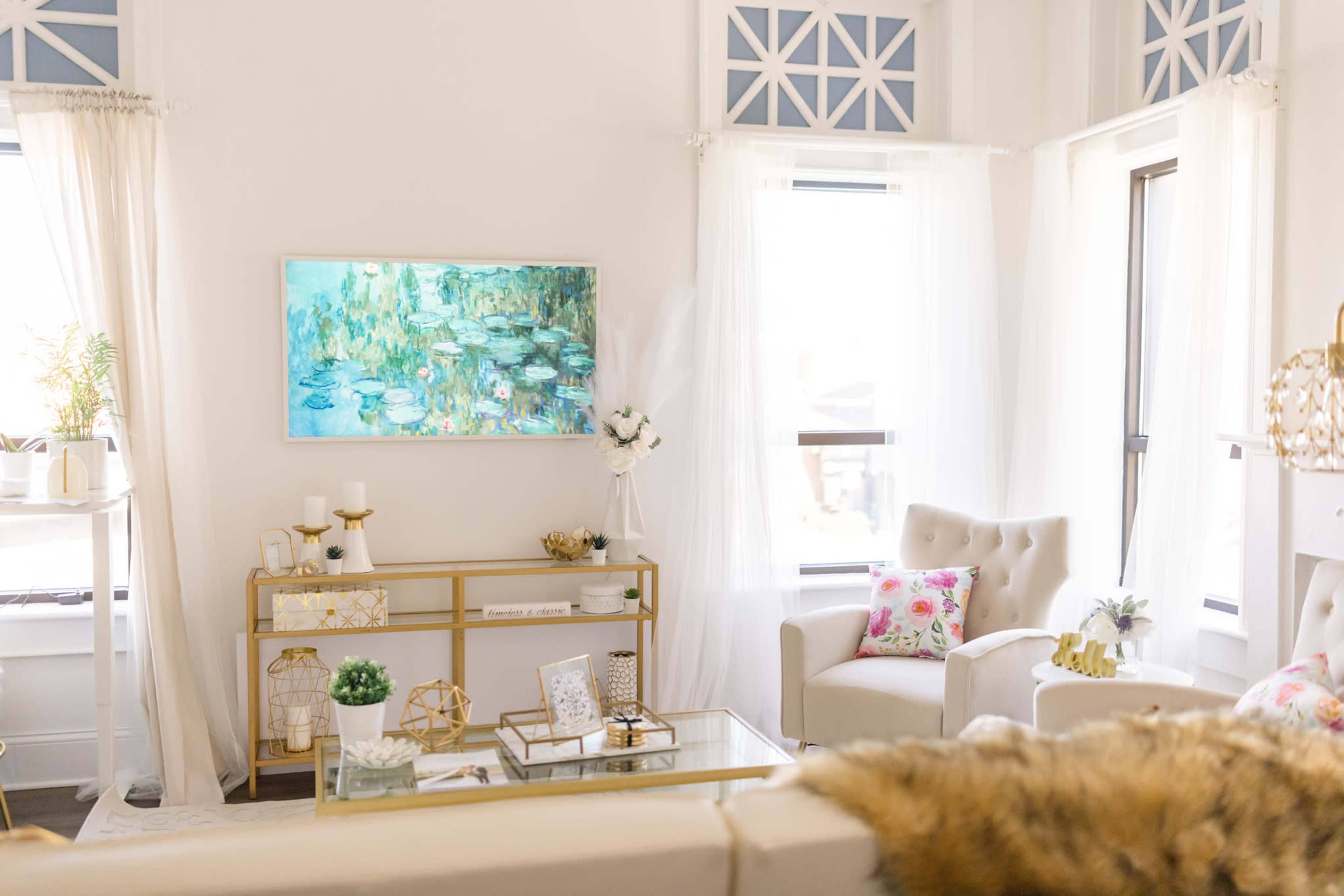 A bright, modern living room with light-colored furniture, a colorful painting on the wall, and decorative elements like plants and pillows.