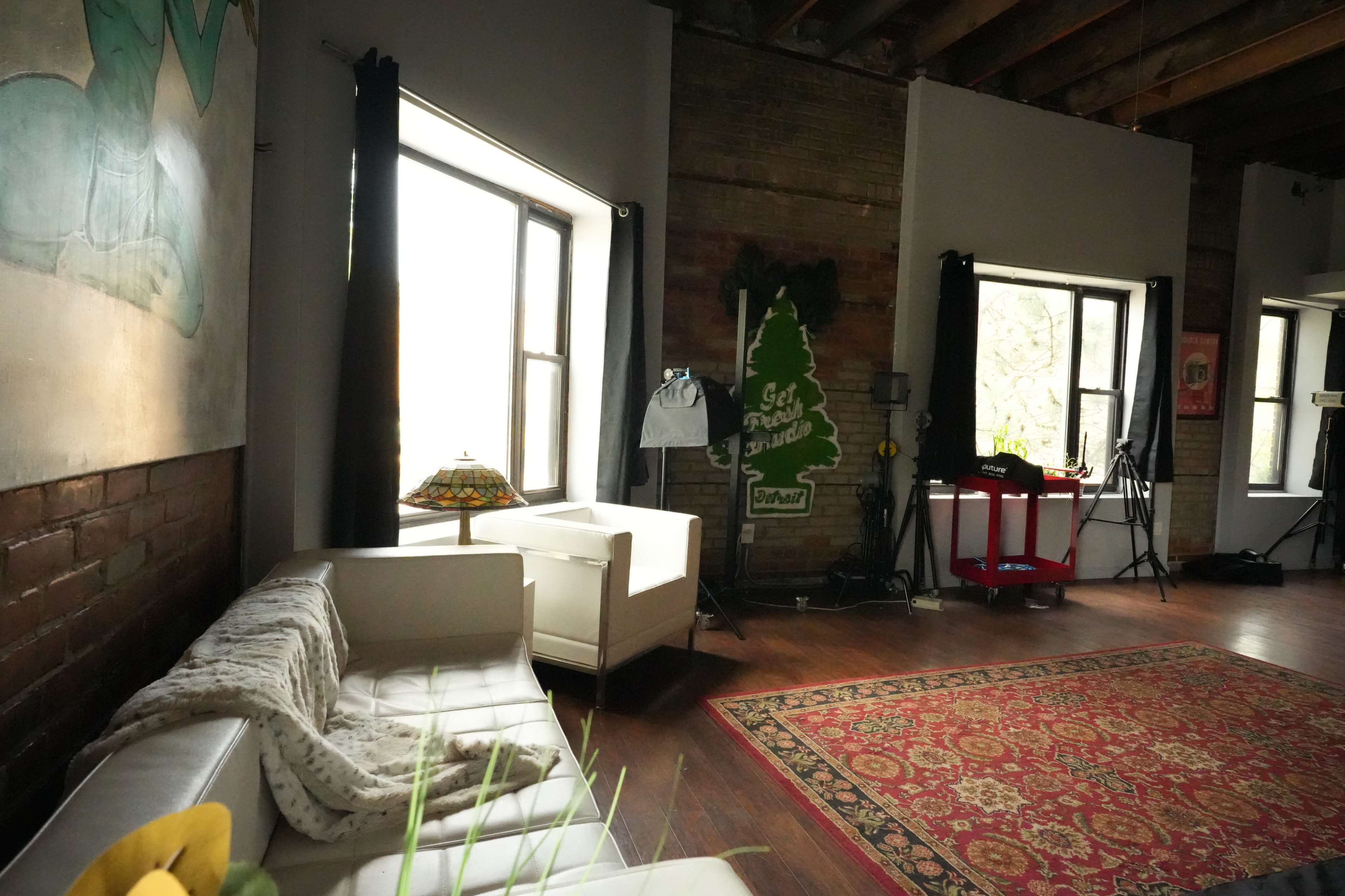 A spacious room with large windows, a white leather sofa, a patterned rug, and various lighting equipment in the background.